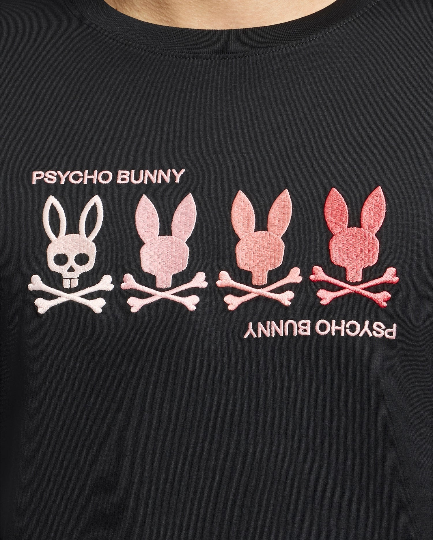 Close-up of the Psycho Bunny Men's Oceania Embroidered Graphic Tee (B6U607F200) featuring four bunny skulls with crossbones in white and pink. "Psycho Bunny" is stitched above and below, creating a unique retro vibe.