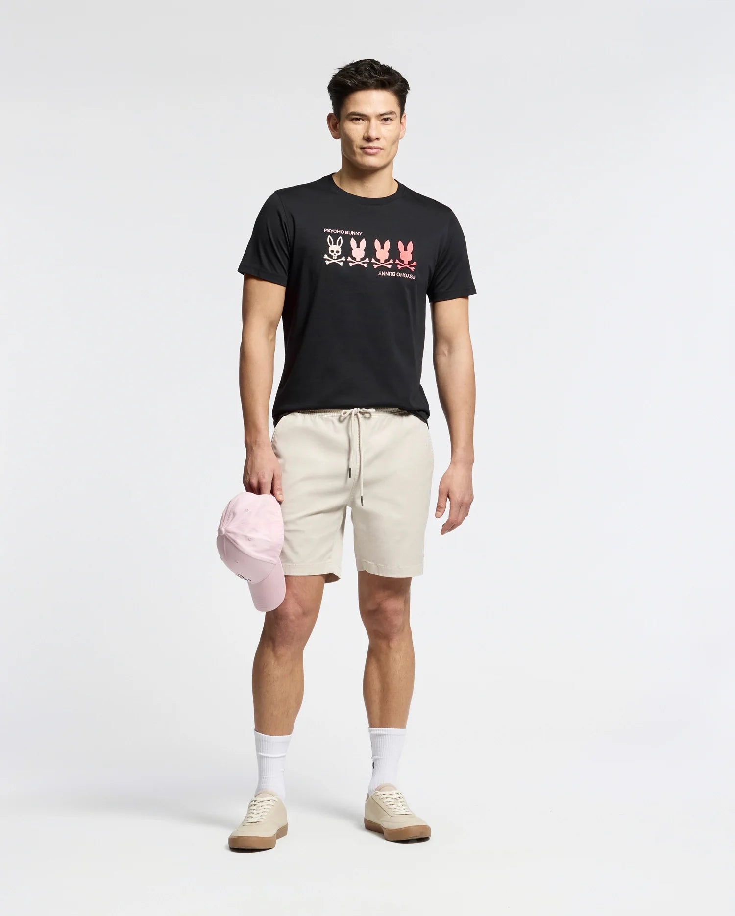 A man stands wearing a Psycho Bunny MENS OCEANIA EMBROIDERED GRAPHIC TEE - B6U607F200, giving off retro vibes. He pairs it with beige shorts, white socks, matching sneakers, and holds a pink cap in his right hand against a plain backdrop.