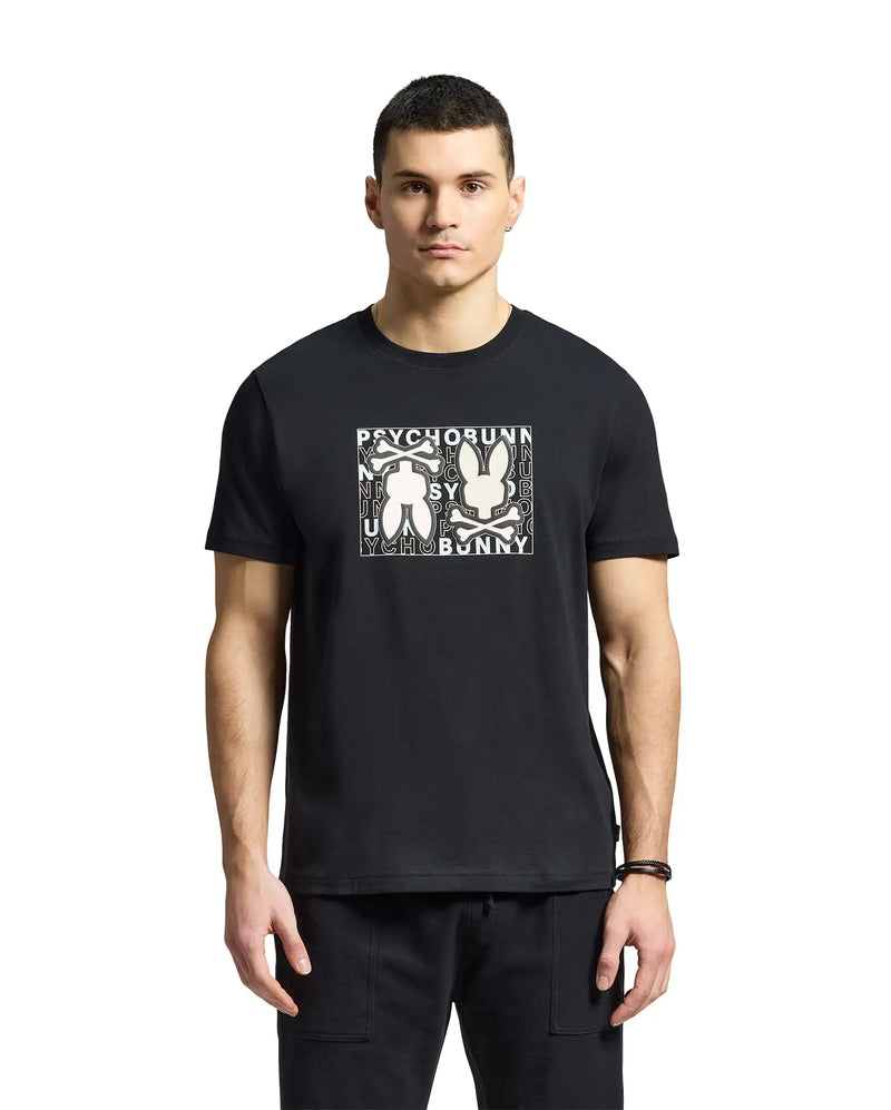 A man is wearing a Psycho Bunny "MENS GOLD COAST GRAPHIC TEE - B6U631F200" in black, made from premium Pima cotton. The classic fit shirt features a graphic bunny design, contrasting with a plain white background.