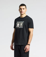 A man in a Psycho Bunny MENS GOLD COAST GRAPHIC TEE (B6U631F200) of premium Pima cotton with abstract figures and text stands against a plain backdrop, looking at the camera. His short hair suits his classic black pants and wristband.