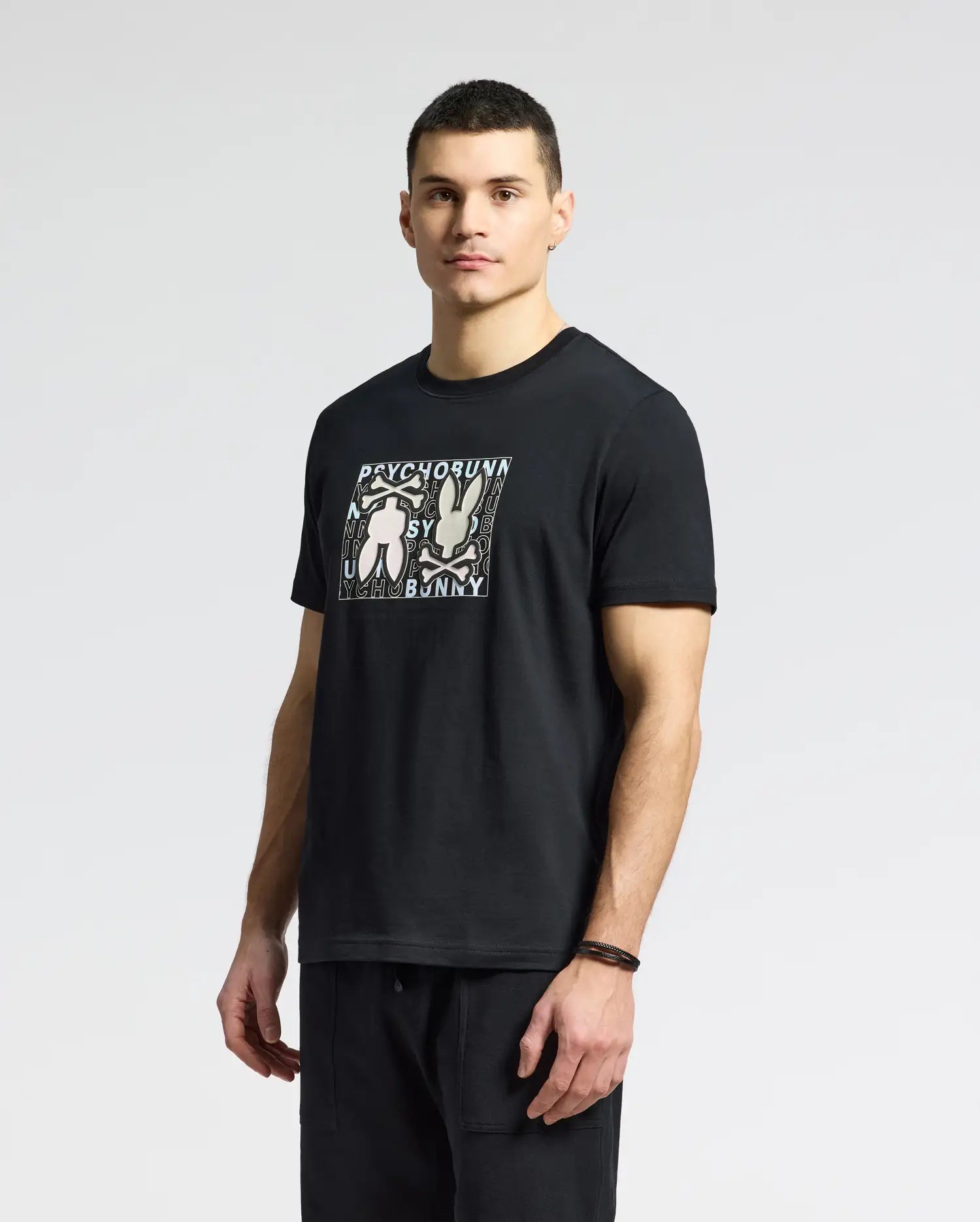 A man in a Psycho Bunny MENS GOLD COAST GRAPHIC TEE (B6U631F200) of premium Pima cotton with abstract figures and text stands against a plain backdrop, looking at the camera. His short hair suits his classic black pants and wristband.