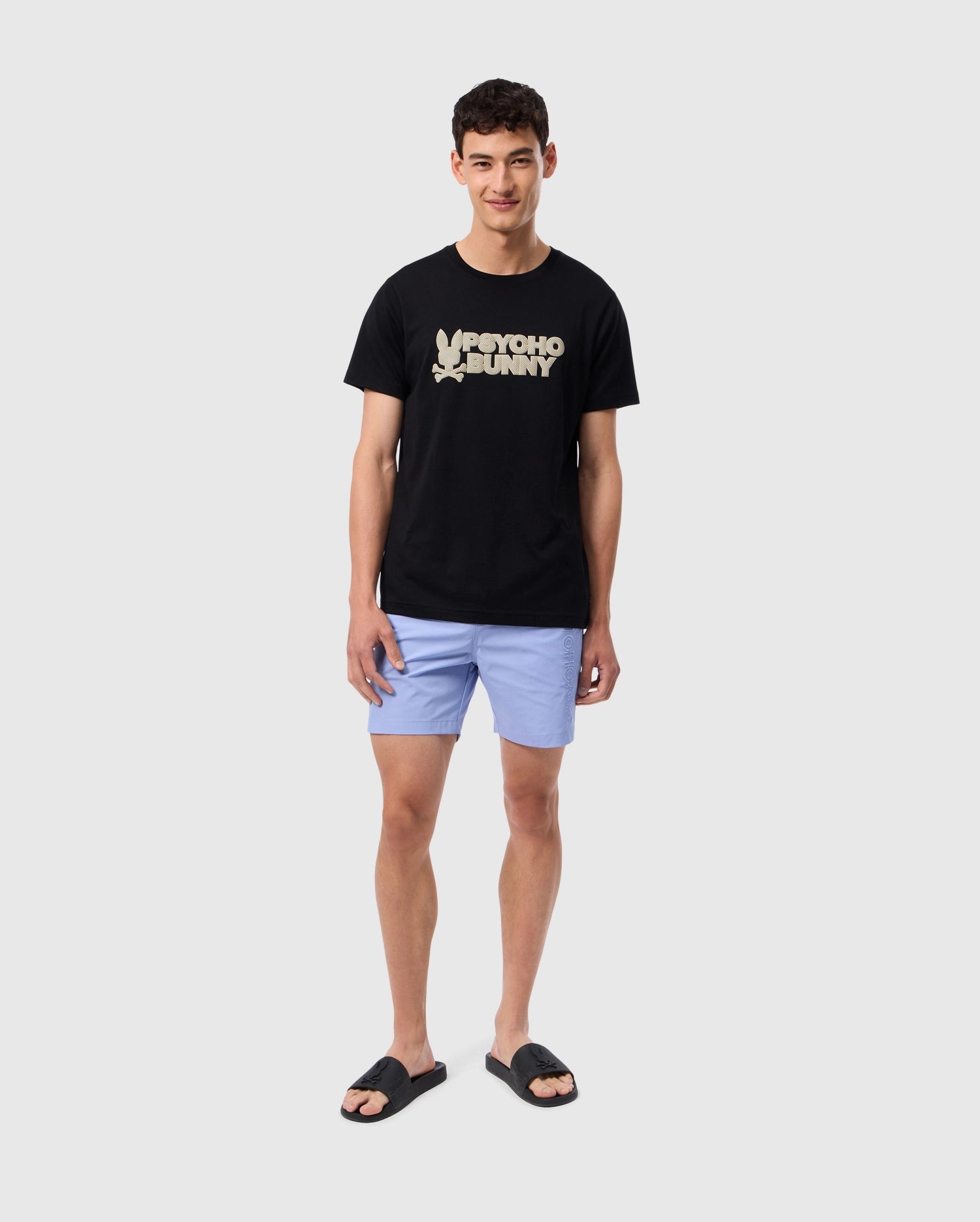 A young man with short dark hair is wearing the Psycho Bunny MENS MELVIN GRAPHIC TEE - B6U663C200 in black. The soft Pima cotton t-shirt showcases an HD-silicone print wordmark graphic. He pairs it with light blue shorts and black sandals, standing against a plain white background, smiling with his arms relaxed by his sides.