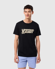 A person with short dark hair is wearing the MENS MELVIN GRAPHIC TEE (B6U663C200) from Psycho Bunny, a black Pima cotton T-shirt featuring white text that reads "Psycho Bunny" accompanied by a stylized bunny logo in HD-silicone print. They are also wearing light blue shorts and standing against a plain light grey background, smiling and looking at the camera.