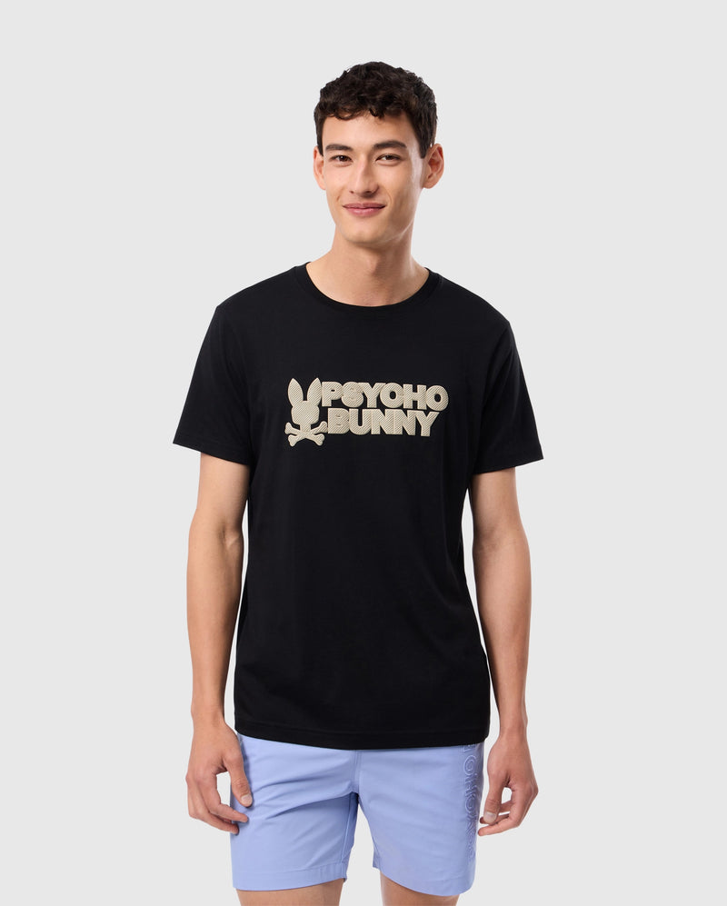 A person with short dark hair is wearing the MENS MELVIN GRAPHIC TEE (B6U663C200) from Psycho Bunny, a black Pima cotton T-shirt featuring white text that reads "Psycho Bunny" accompanied by a stylized bunny logo in HD-silicone print. They are also wearing light blue shorts and standing against a plain light grey background, smiling and looking at the camera.
