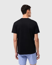 A person with short, dark hair stands with their back to the camera, wearing a black Psycho Bunny MENS MELVIN GRAPHIC TEE - B6U663C200 featuring an HD-silicone print and light blue shorts. The background is plain and light gray.