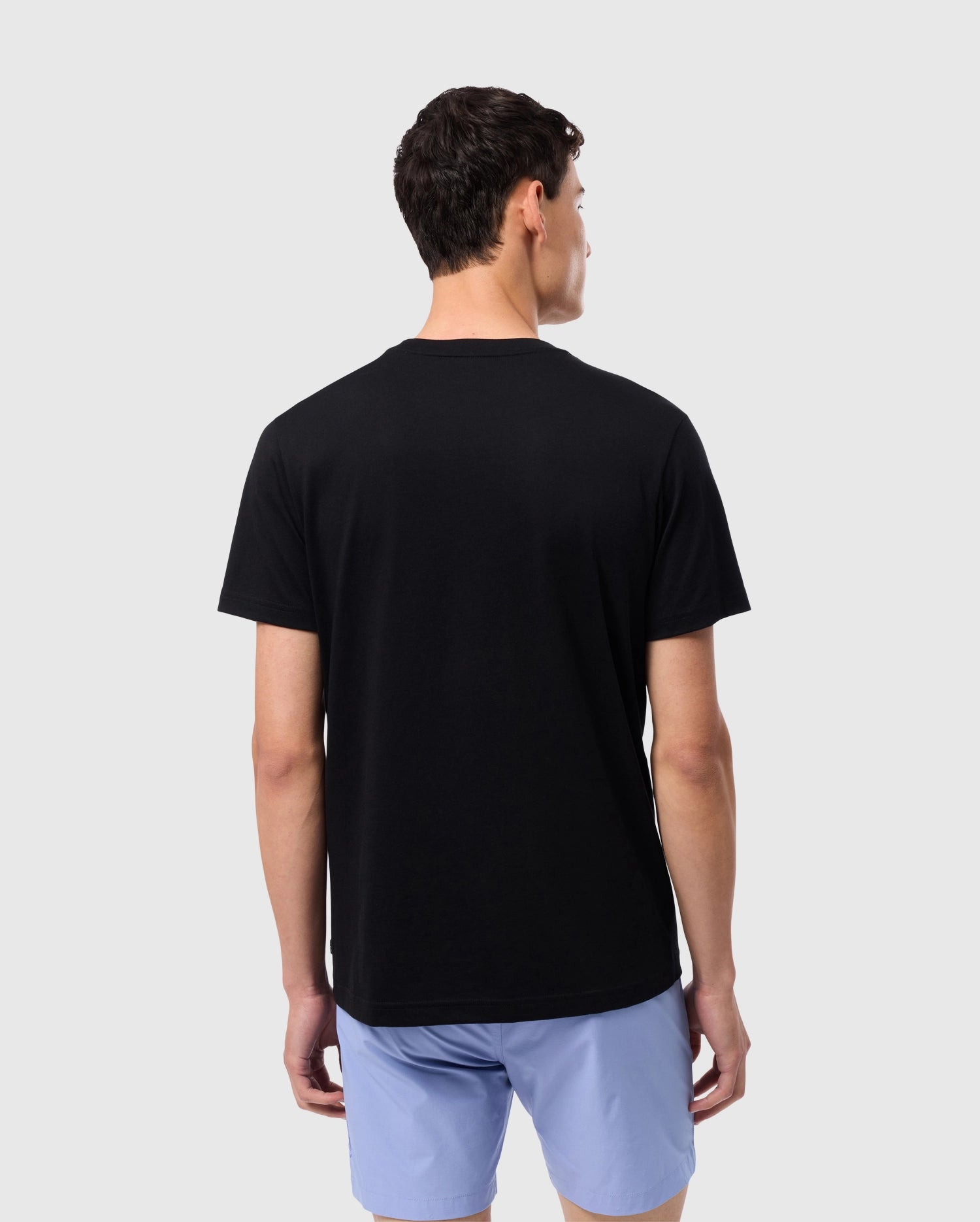 A person with short, dark hair stands with their back to the camera, wearing a black Psycho Bunny MENS MELVIN GRAPHIC TEE - B6U663C200 featuring an HD-silicone print and light blue shorts. The background is plain and light gray.