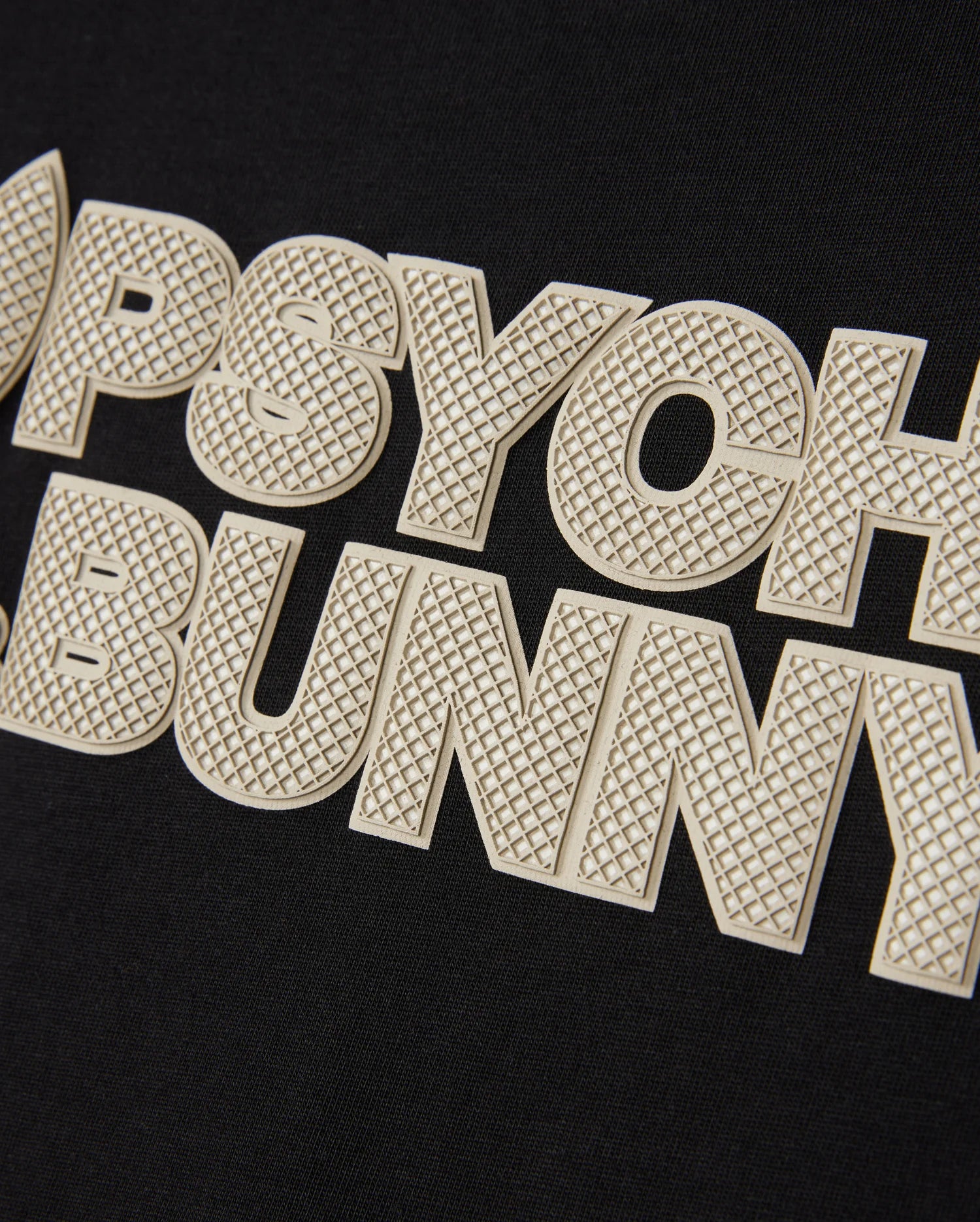 A close-up shot of the textured text "PSYCHO BUNNY" in large, bold, off-white letters on a black background. The wordmark graphic features a fine, grid-like pattern on the letters, providing a tactile, embossed look reminiscent of HD-silicone print. This detail is featured on the Psycho Bunny MENS MELVIN GRAPHIC TEE - B6U663C200.