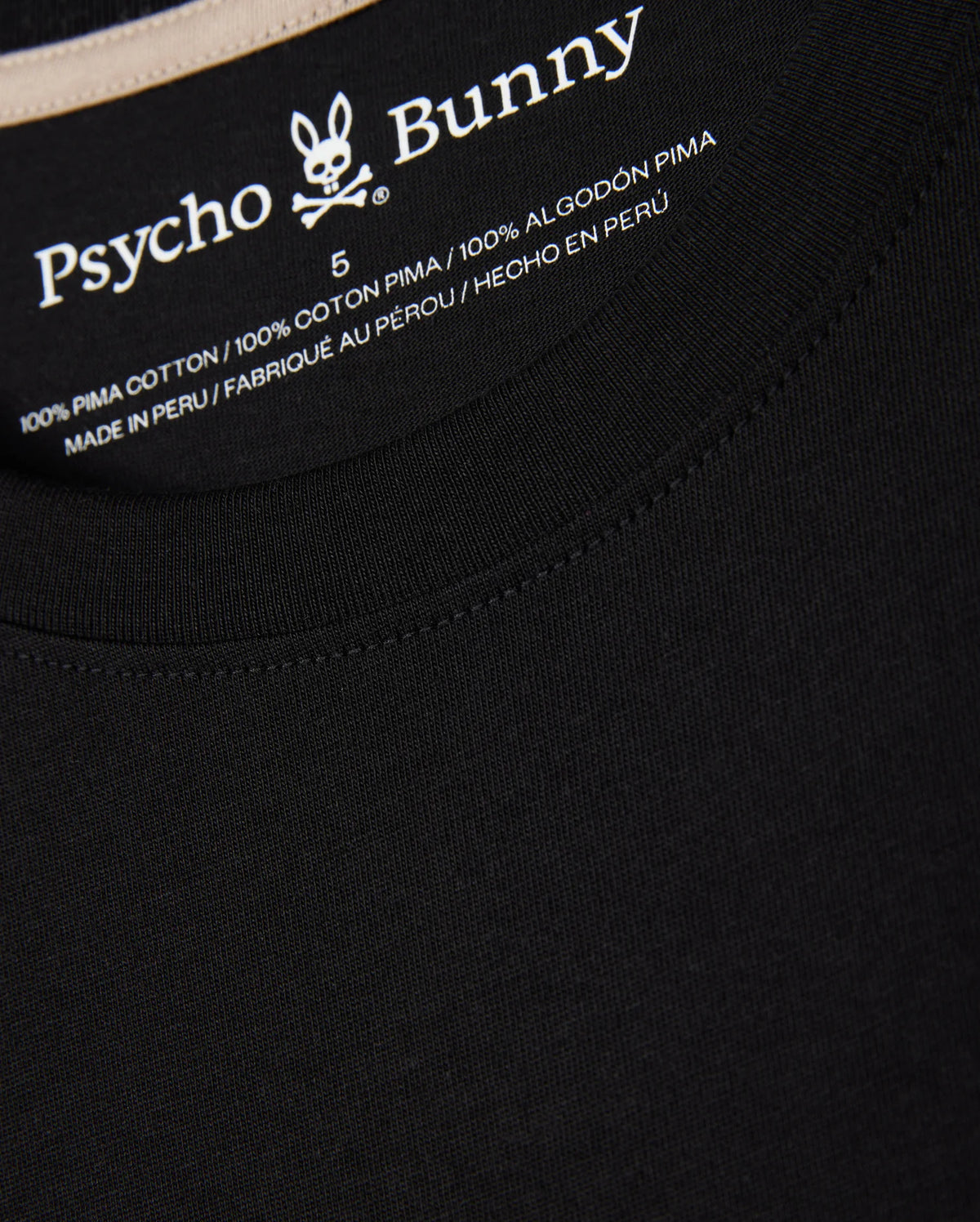 Close-up of the neck tag on the MENS MELVIN GRAPHIC TEE - B6U663C200 black T-shirt displaying the Psycho Bunny logo and wordmark graphic along with size 5. The T-shirt is made of 100% Pima cotton and manufactured in Peru, with text in English, French, and Spanish.