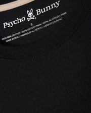Close-up of the neck tag on the MENS MELVIN GRAPHIC TEE - B6U663C200 black T-shirt displaying the Psycho Bunny logo and wordmark graphic along with size 5. The T-shirt is made of 100% Pima cotton and manufactured in Peru, with text in English, French, and Spanish.