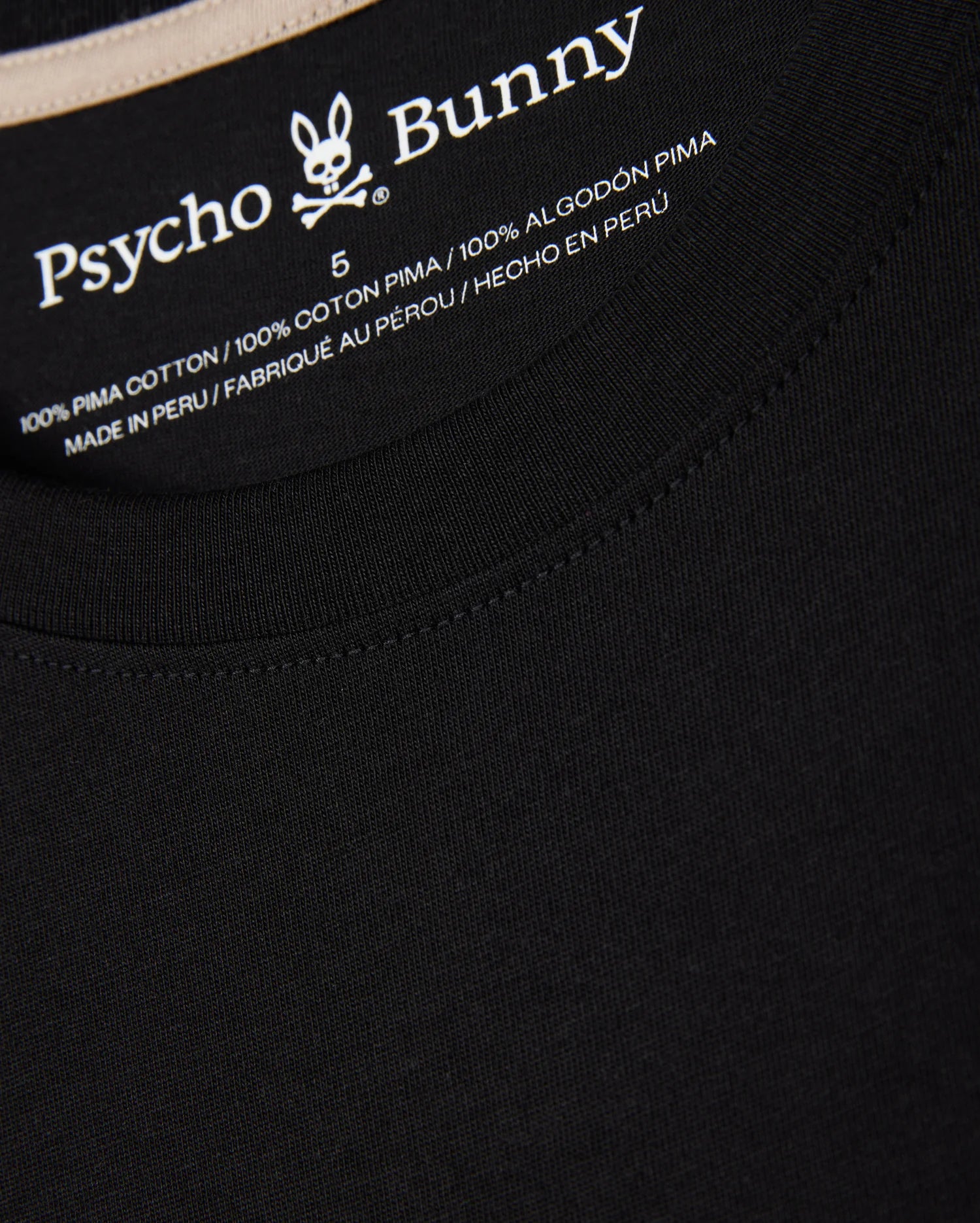 Close-up of the neck tag on the MENS MELVIN GRAPHIC TEE - B6U663C200 black T-shirt displaying the Psycho Bunny logo and wordmark graphic along with size 5. The T-shirt is made of 100% Pima cotton and manufactured in Peru, with text in English, French, and Spanish.