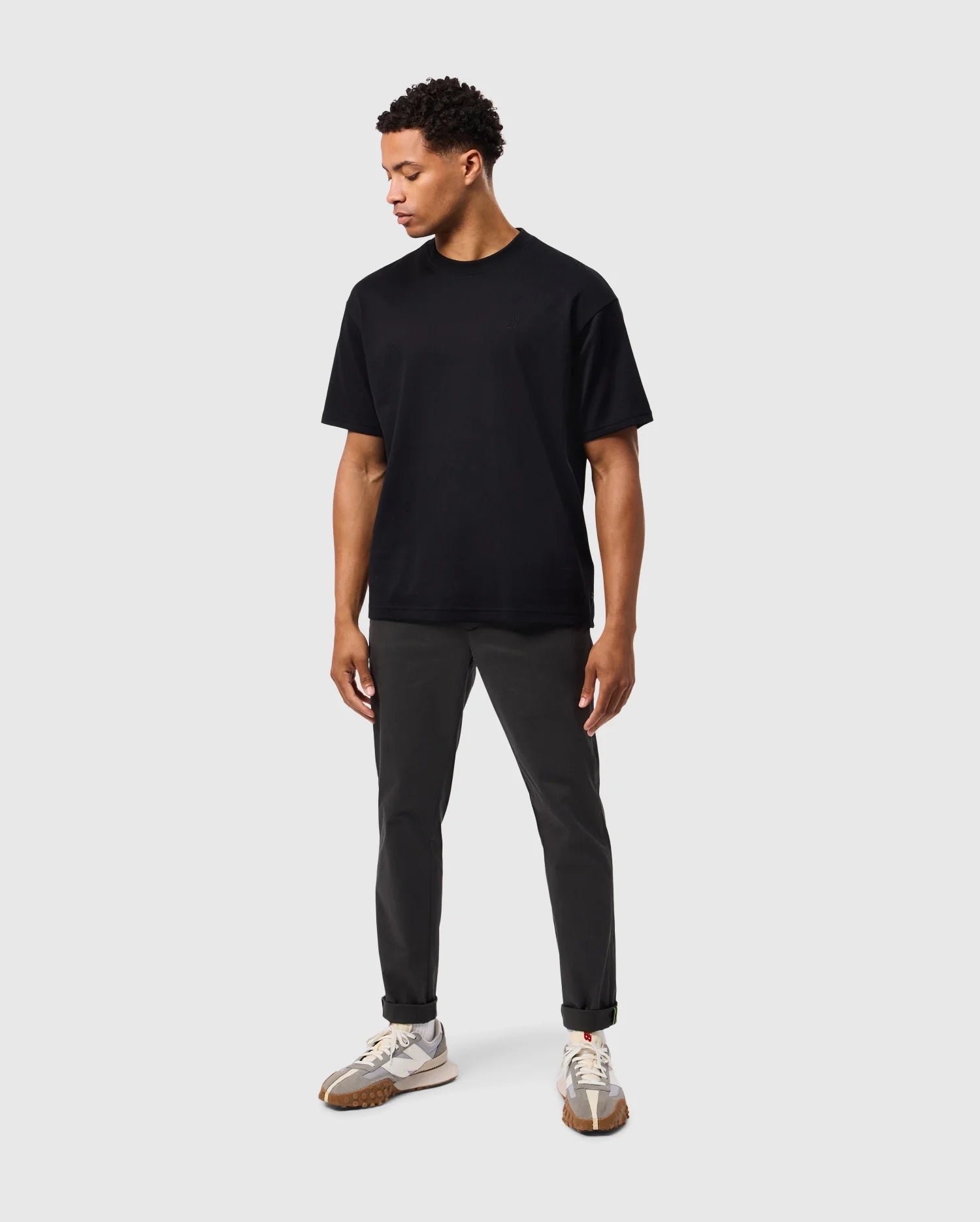 A person is standing against a plain grey background, wearing the MENS DUKE OVERSIZED TEE - B6U742D200 by Psycho Bunny, dark grey pants, and white sneakers. The relaxed fit t-shirt hangs comfortably as they look downward to their right with their arms by their sides.