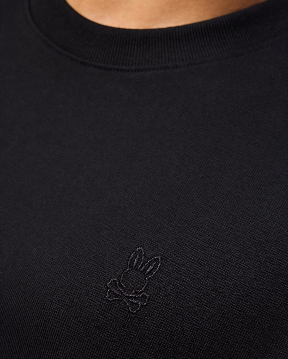 Close-up of a person wearing a Psycho Bunny MENS DUKE OVERSIZED TEE - B6U742D200, crafted from black heavyweight Pima cotton with a ribbed crew neckline. The relaxed fit tee features a subtle black embroidered chest bunny head with crossbones. The background is out of focus.