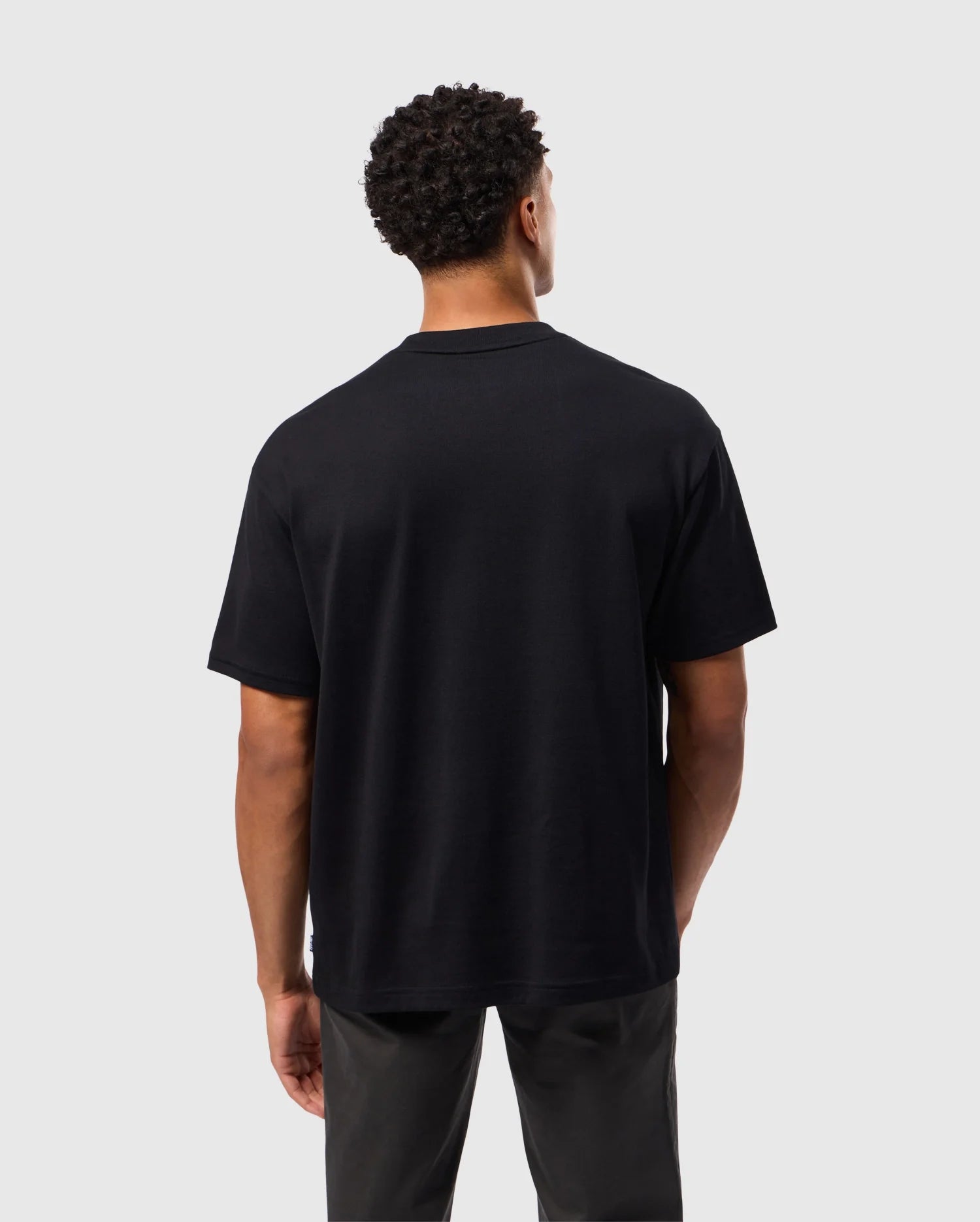 A person with short, curly hair is standing with their back to the camera. They are wearing a relaxed fit MENS DUKE OVERSIZED TEE - B6U742D200 by Psycho Bunny, made of heavyweight Pima cotton, paired with dark pants. The background is plain and light grey.