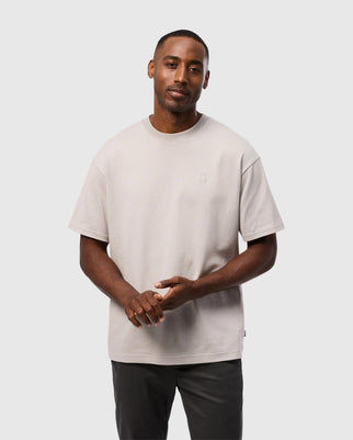 A man with short hair and a trimmed beard stands against a plain background. He wears the Psychobunny MEN'S DUKE OVERSIZED TEE - B6U742D200, a light gray, relaxed-fit shirt made of heavyweight Pima cotton, featuring a small embroidered chest bunny logo. This is paired with dark green pants. His hands are clasped in front of him as he looks into the camera with a neutral expression.