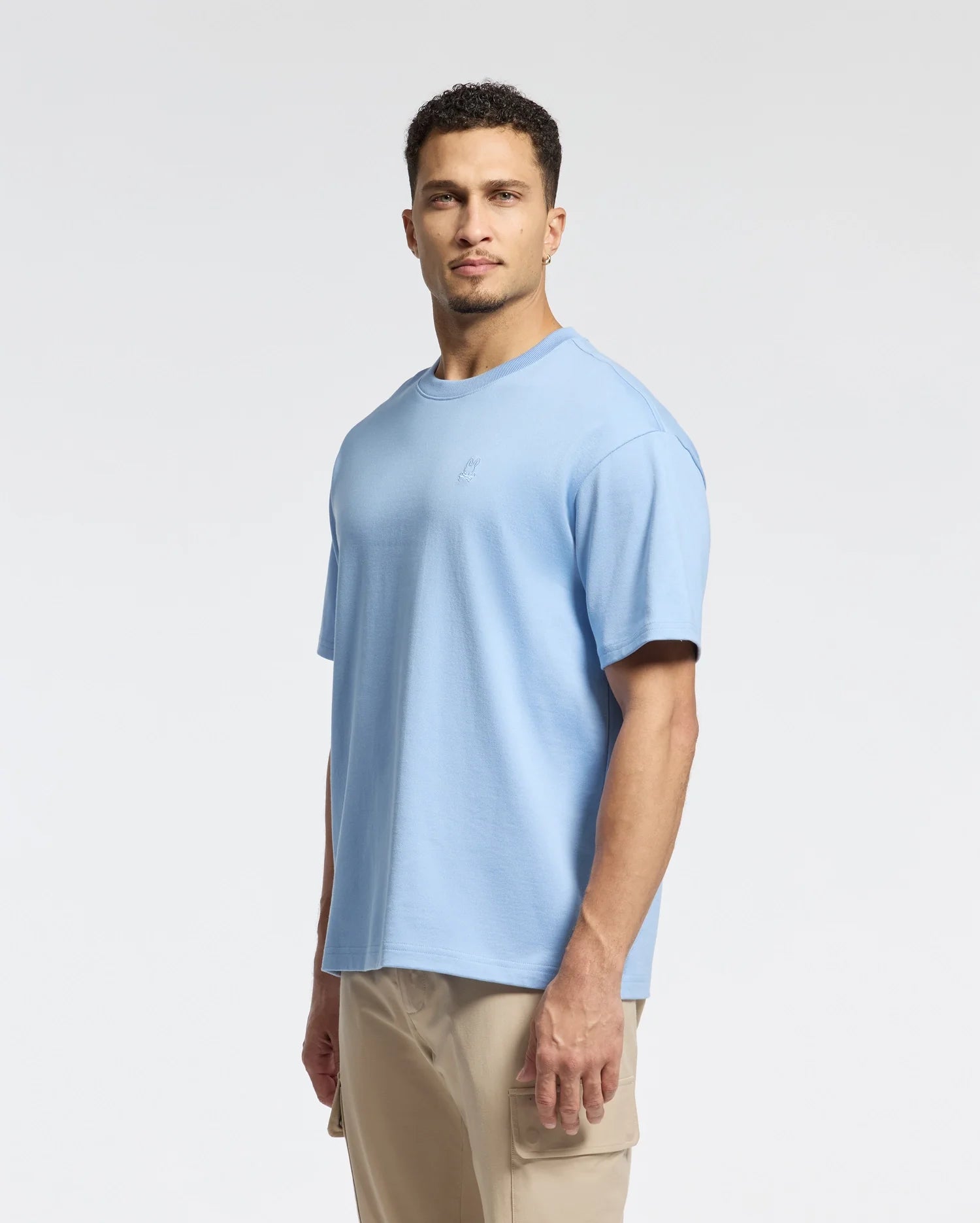 A man models the Psycho Bunny Men's Duke Heavy Weight Relaxed Fit Tee (B6U742D200) in light blue with beige pants against a plain white background. His expression is neutral, facing forward with his body slightly angled, highlighting the tee's relaxed fit.