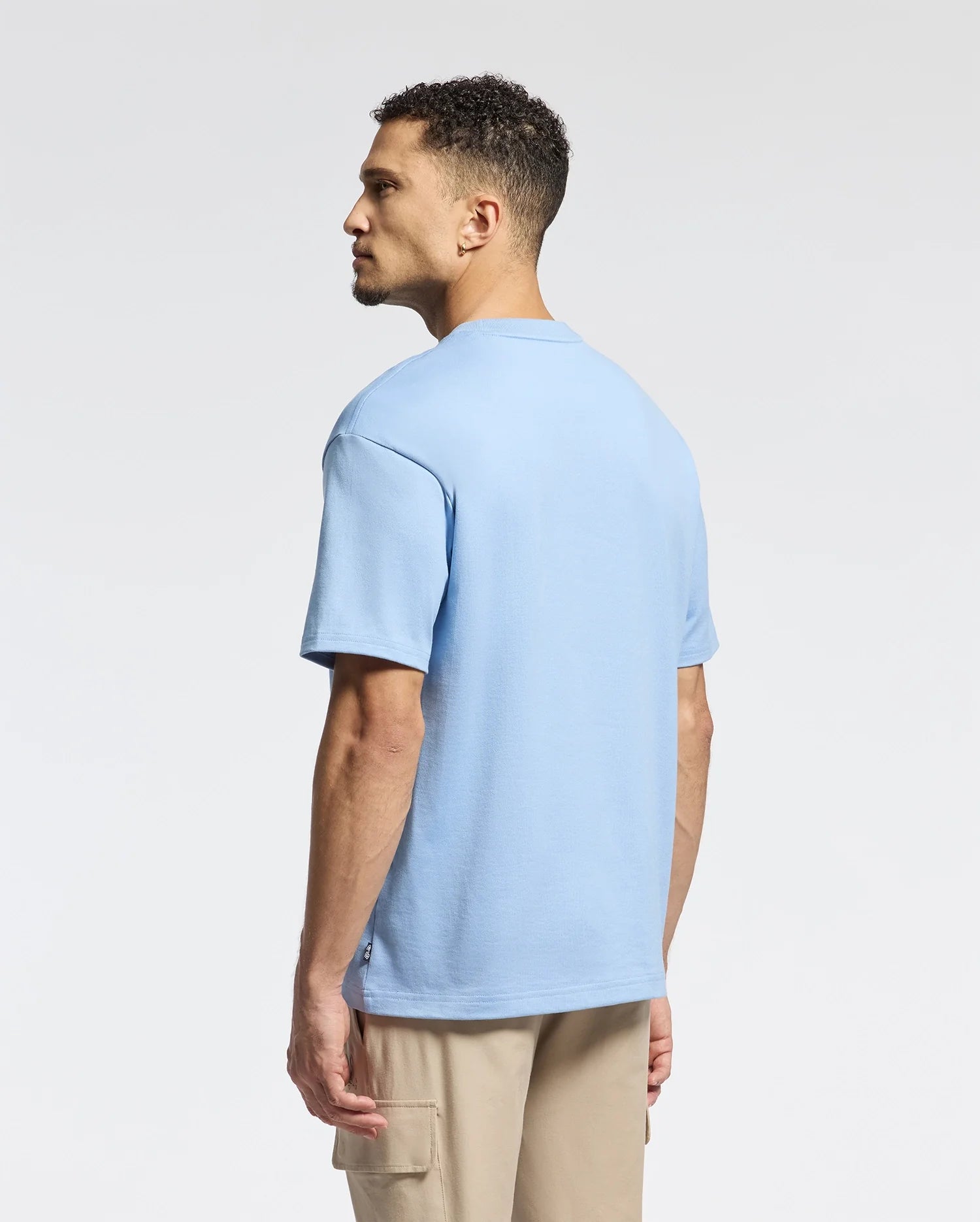 A man with short curly hair wears a Psycho Bunny MENS DUKE HEAVY WEIGHT RELAXED FIT TEE - B6U742D200 in light blue and beige cargo pants. He stands with his back to the camera, head turned sideways, against a neutral light gray background.