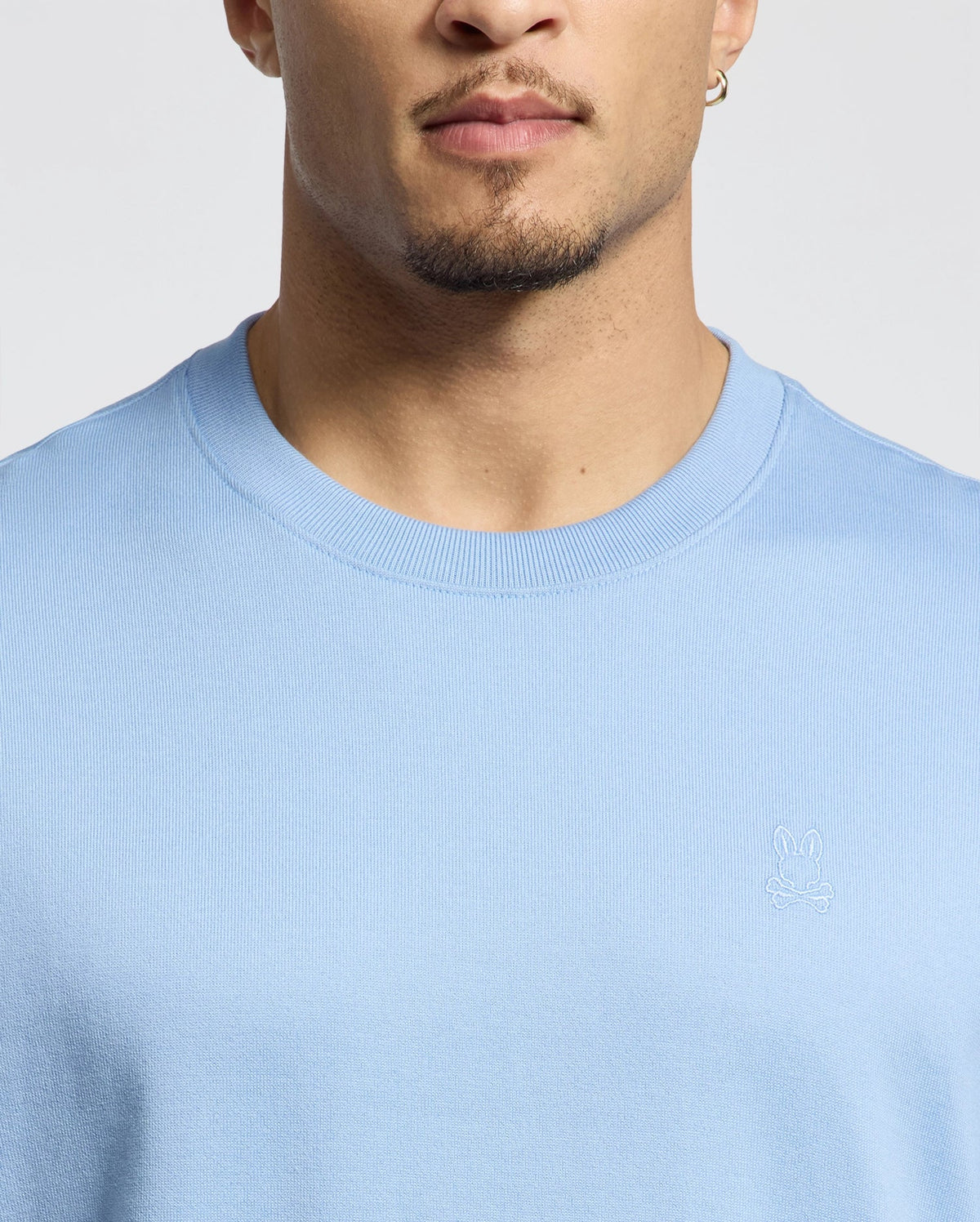 A person in a light blue Psycho Bunny MENS DUKE HEAVY WEIGHT RELAXED FIT TEE with a small embroidered rabbit logo, made from luxurious Pima cotton. They have short hair, a light beard, and wear a small hoop earring, showing the tee's relaxed fit and their neutral facial expression.