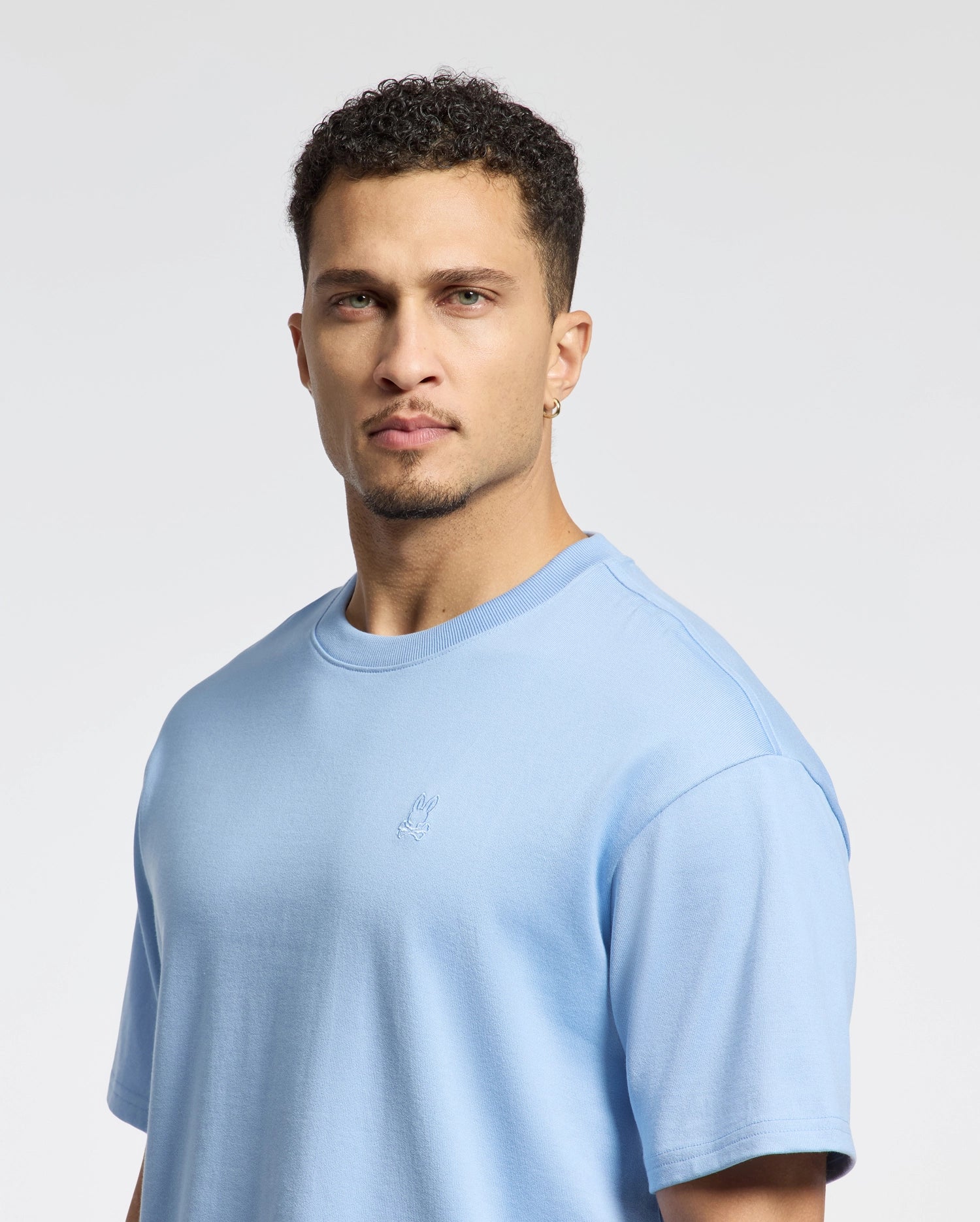A man with short curly hair and a goatee wears a Psycho Bunny light blue heavyweight jersey T-shirt (MENS DUKE HEAVY WEIGHT RELAXED FIT TEE - B6U742D200) featuring a small chest embroidery. He has an earring in his left ear, looks directly at the camera with a neutral expression, and stands before a plain background.
