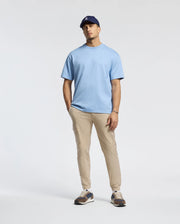 A person in a Psycho Bunny Duke Heavy Weight Relaxed Fit Tee, light blue, paired with beige pants and a navy cap, poses against a plain background with one hand in their pocket and wearing sneakers.