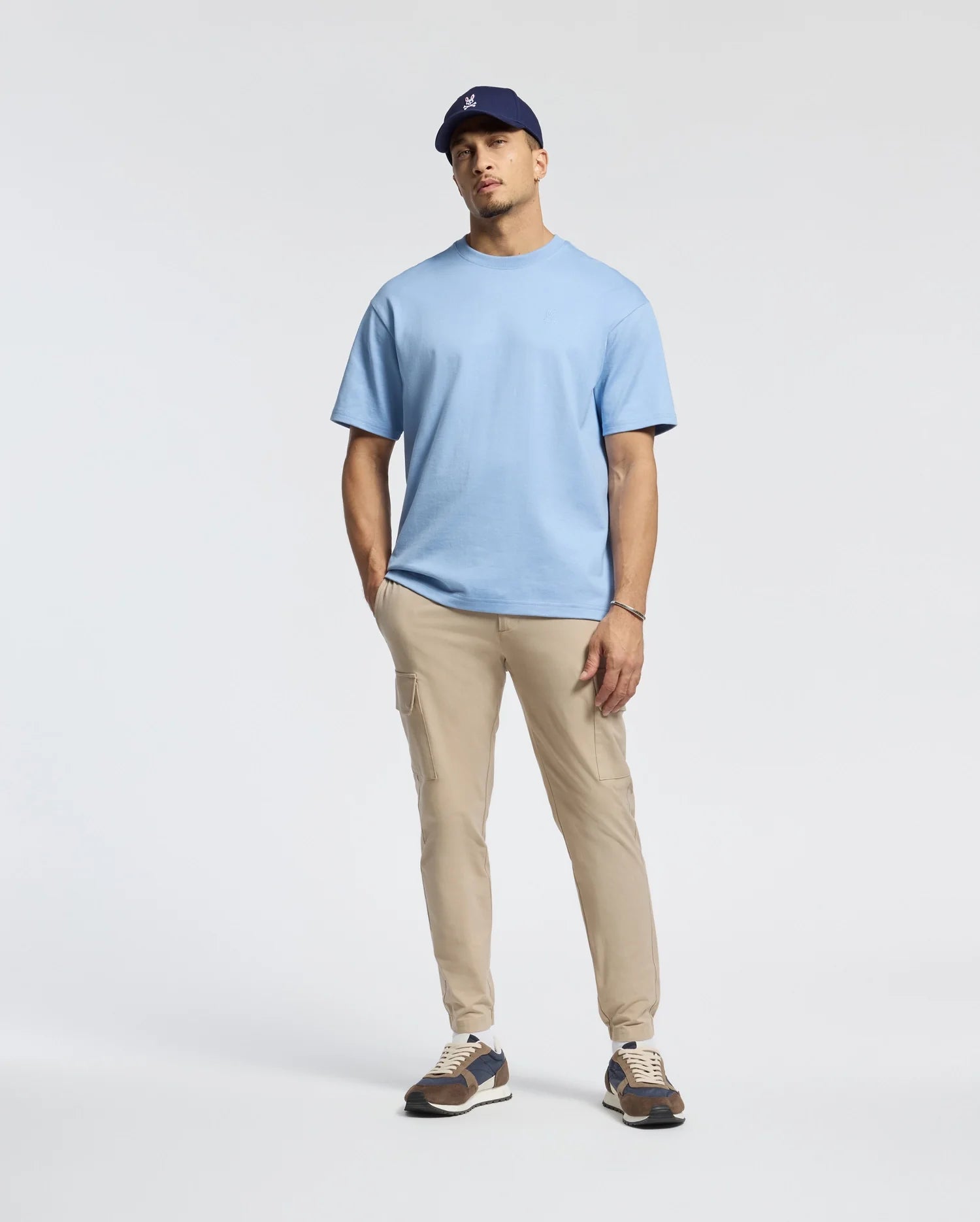 A person in a Psycho Bunny Duke Heavy Weight Relaxed Fit Tee, light blue, paired with beige pants and a navy cap, poses against a plain background with one hand in their pocket and wearing sneakers.