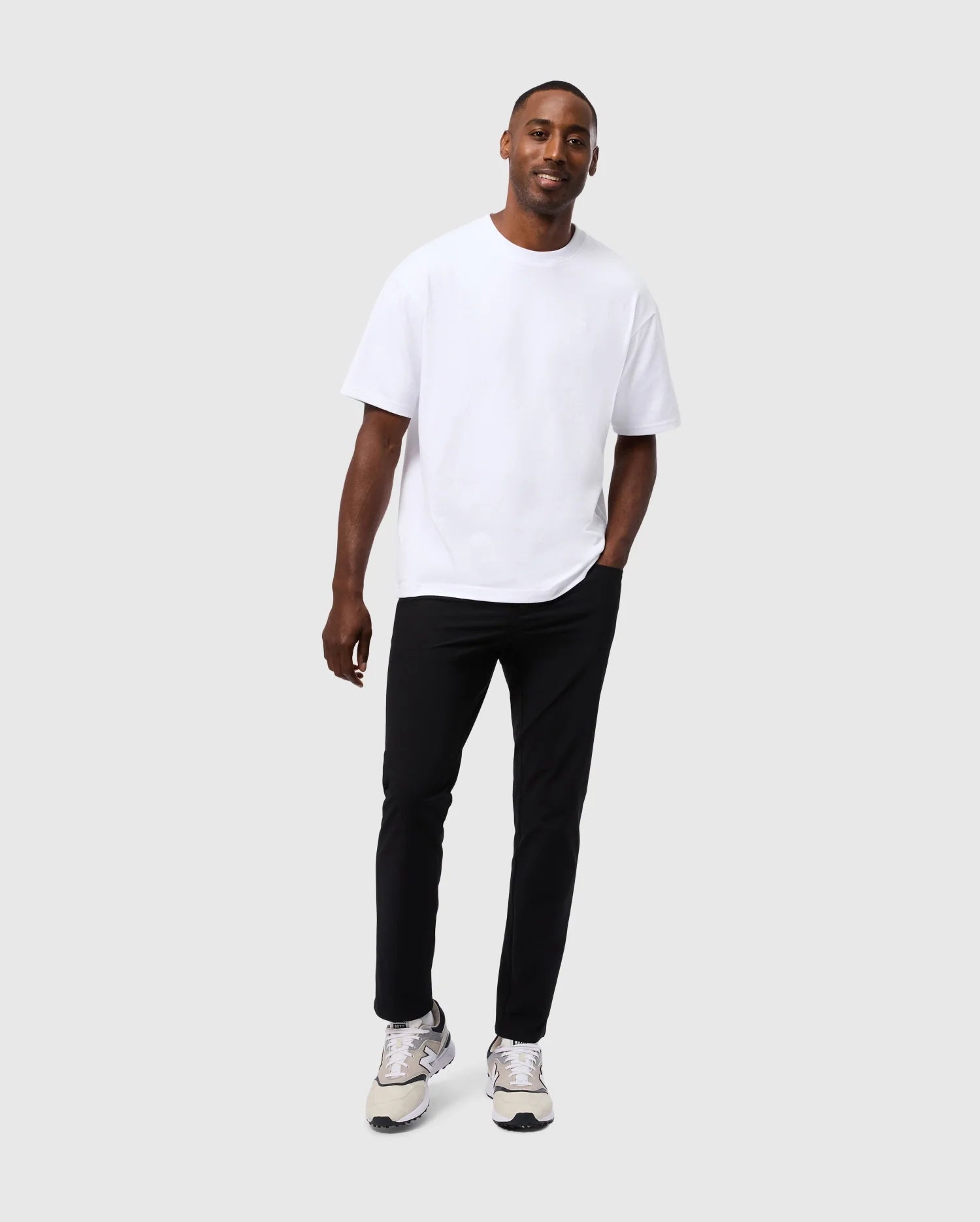 A man stands against a plain background, smiling and wearing the Psycho Bunny MENS DUKE OVERSIZED TEE - B6U742D200 made of heavyweight Pima cotton, black pants, and white sneakers. One hand is in his pocket while he appears relaxed and casual.