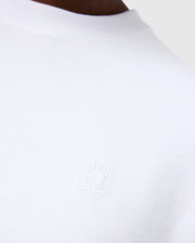 Close-up of a person wearing the Psycho Bunny MENS DUKE OVERSIZED TEE - B6U742D200, crafted from heavyweight Pima cotton in a relaxed fit. The left chest showcases an embroidered white bunny with crossbones beneath it. The background is neutral and minimalistic.