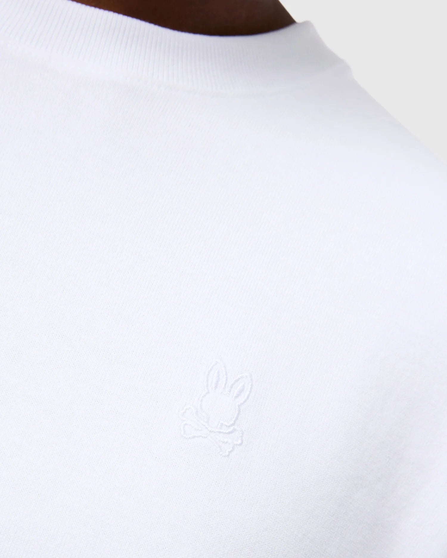Close-up of a person wearing the Psycho Bunny MENS DUKE OVERSIZED TEE - B6U742D200, crafted from heavyweight Pima cotton in a relaxed fit. The left chest showcases an embroidered white bunny with crossbones beneath it. The background is neutral and minimalistic.
