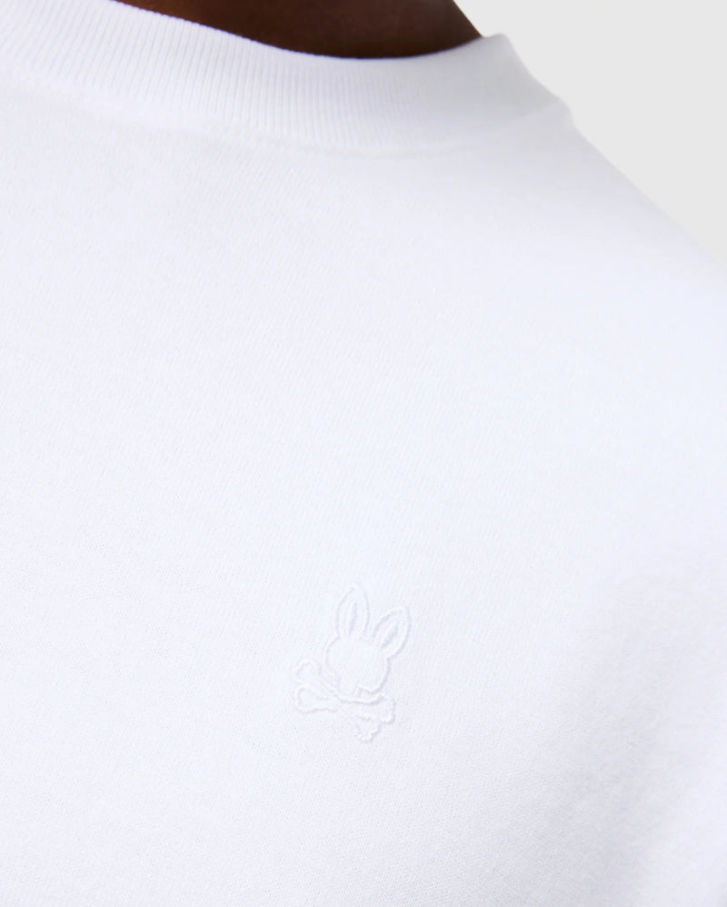 Close-up of a person wearing the Psycho Bunny MENS DUKE OVERSIZED TEE - B6U742D200, crafted from heavyweight Pima cotton in a relaxed fit. The left chest showcases an embroidered white bunny with crossbones beneath it. The background is neutral and minimalistic.