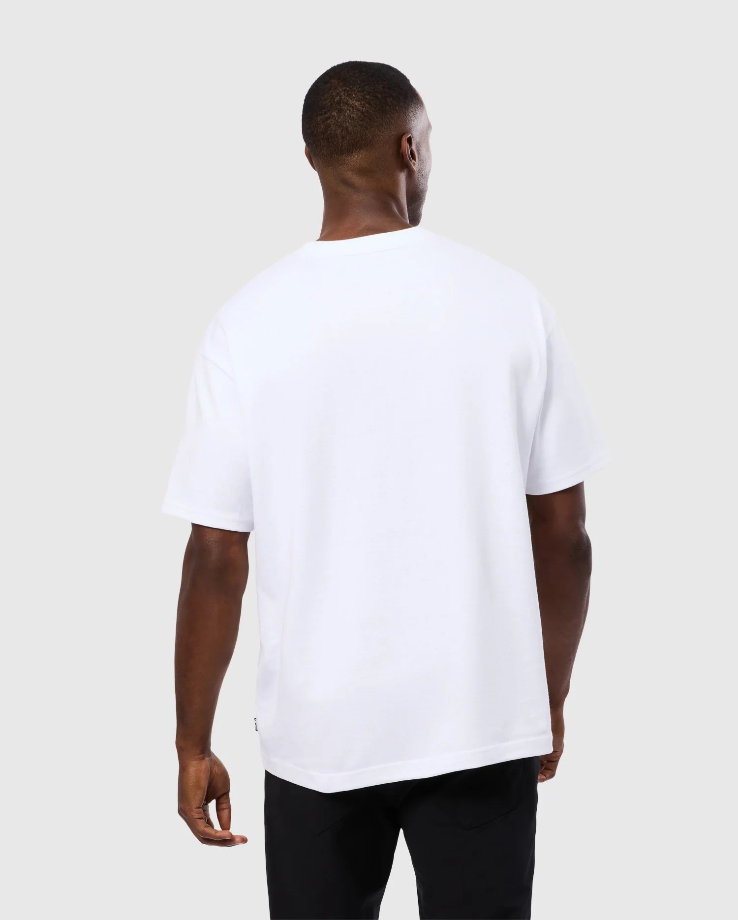 A person with short hair is standing with their back to the camera. They are wearing a plain white MENS DUKE OVERSIZED TEE - B6U742D200 by Psycho Bunny and black pants. The background is light gray.