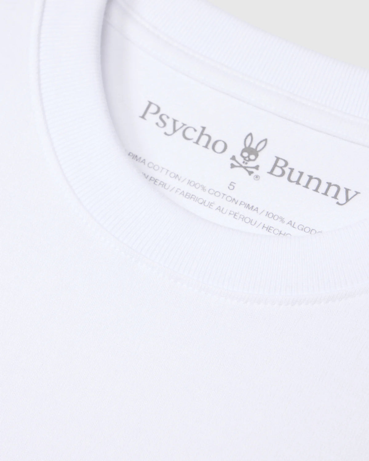 Close-up of a white Psycho Bunny MENS DUKE OVERSIZED TEE - B6U742D200 collar, displaying a printed label with the brand’s typography and logo, featuring a bunny with crossbones. Text indicates the shirt is made from 100% heavyweight Pima cotton and made in Peru.