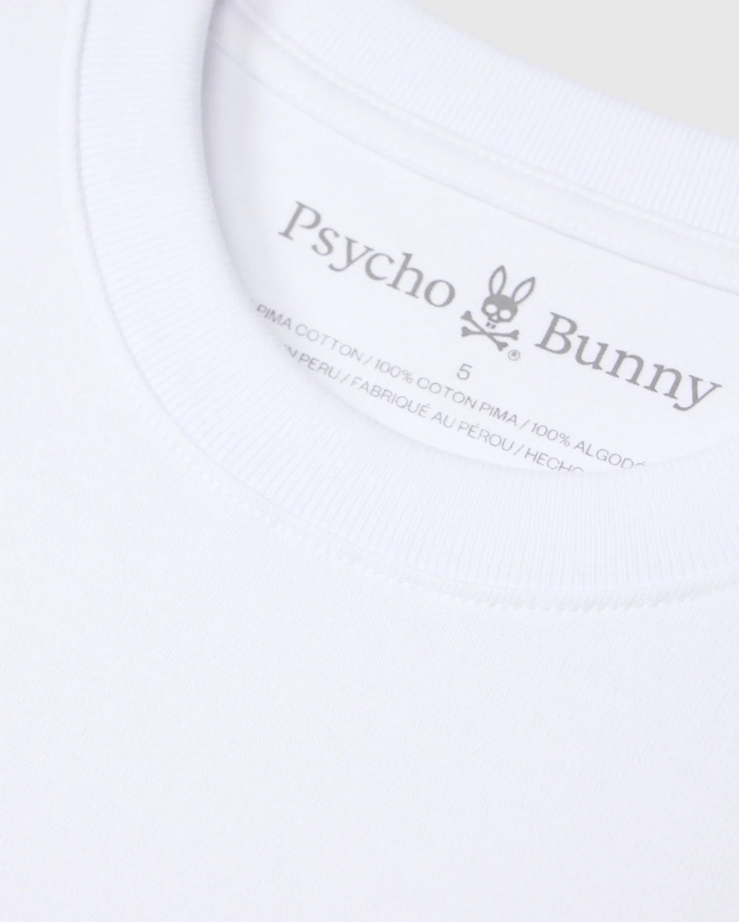 Close-up of a white Psycho Bunny MENS DUKE OVERSIZED TEE - B6U742D200 collar, displaying a printed label with the brand’s typography and logo, featuring a bunny with crossbones. Text indicates the shirt is made from 100% heavyweight Pima cotton and made in Peru.