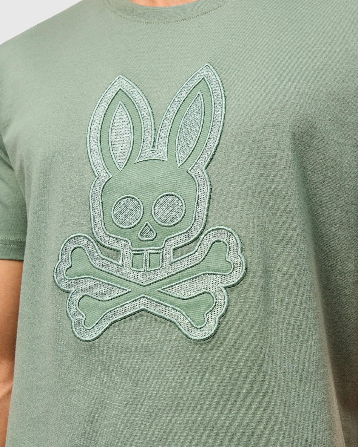 Product Name: MENS MALTON GRAPHIC TEE - B6U795D200
Brand Name: Psycho Bunny

Close-up of a person wearing the Psycho Bunny MENS MALTON GRAPHIC TEE - B6U795D200 in light green Pima cotton, featuring a stylized bunny skull and crossbones design prominently embossed in the center. This graphic tee combines style and comfort with its soft fabric, making it an essential addition to any wardrobe. Made in Peru.