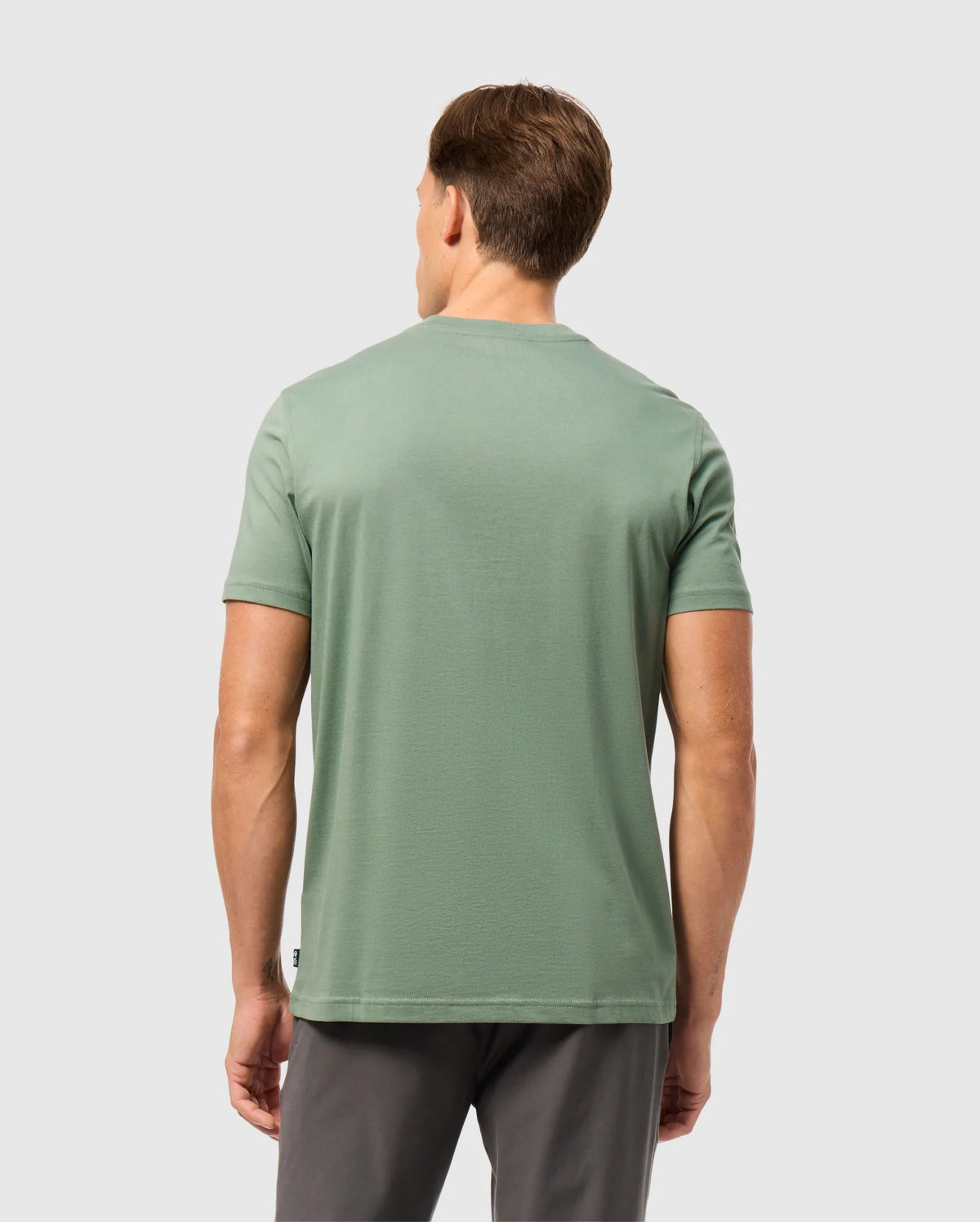 A man with short brown hair is standing with his back to the camera, wearing a green Psycho Bunny MENS MALTON GRAPHIC TEE - B6U795D200 and dark gray pants against a plain light gray background.