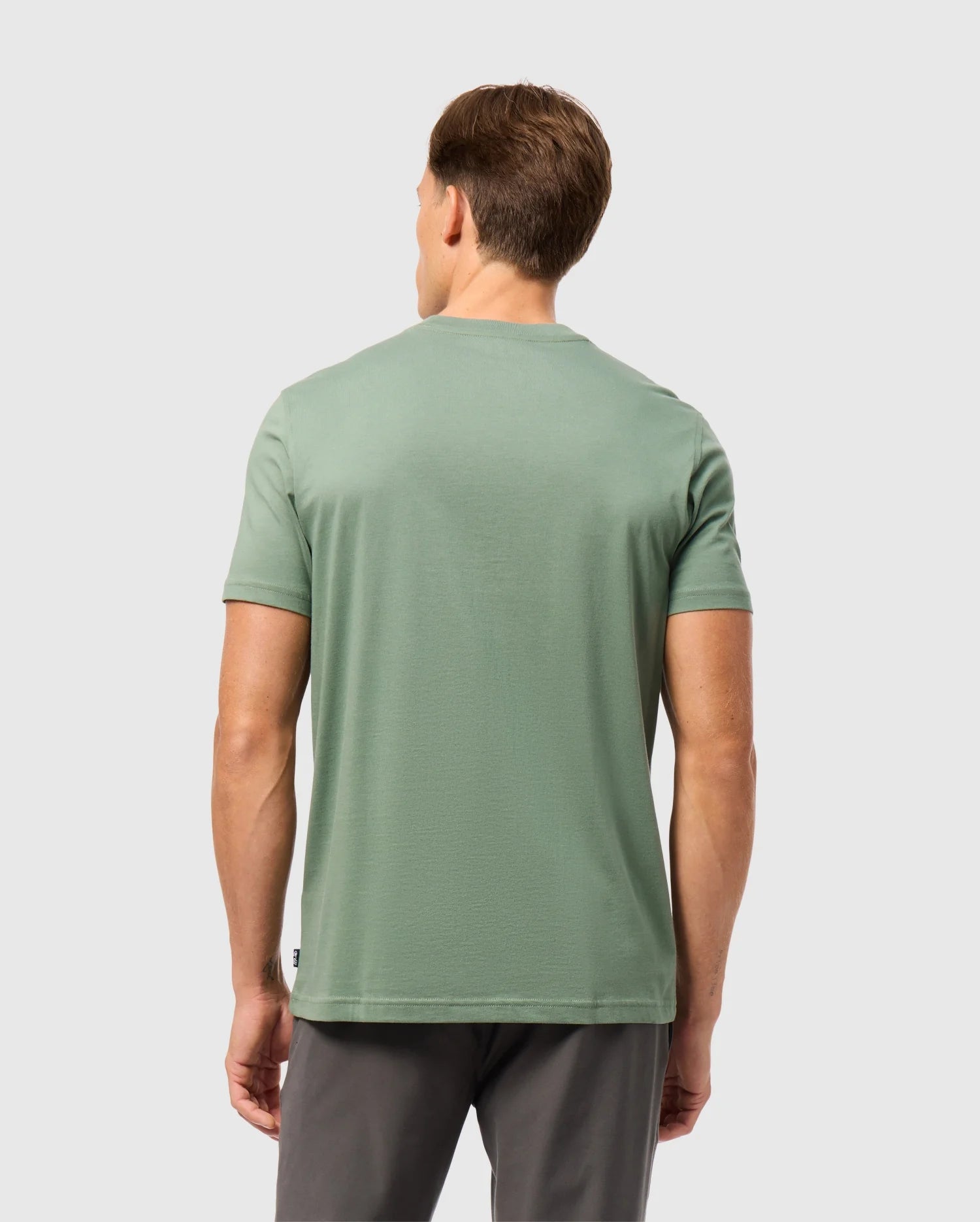 A man with short brown hair is standing with his back to the camera, wearing a green Psycho Bunny MENS MALTON GRAPHIC TEE - B6U795D200 and dark gray pants against a plain light gray background.
