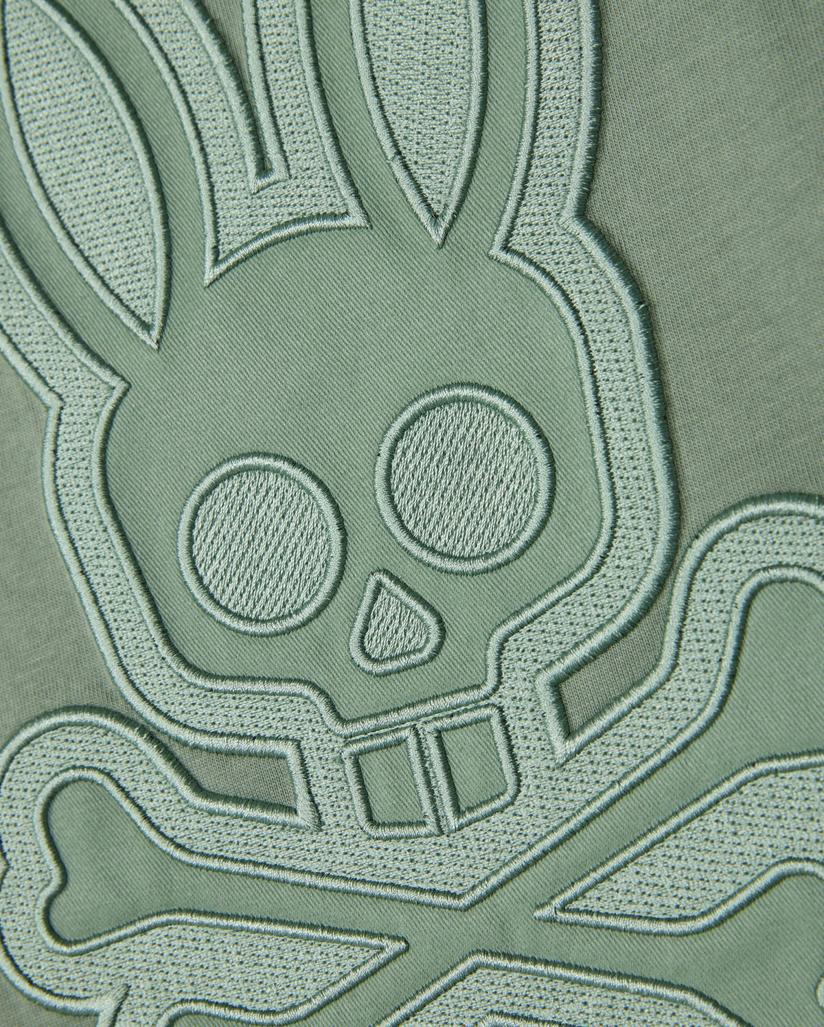 A close-up of an embroidered patch on a Psycho Bunny MENS MALTON GRAPHIC TEE - B6U795D200 made from Pima cotton fabric. The design features a stylized skull with bunny ears and crossbones, detailed with stitching that raises it from the muted green background, giving it a three-dimensional look. Made in Peru, the unique appeal of this T-shirt is enhanced by its intricate craftsmanship.