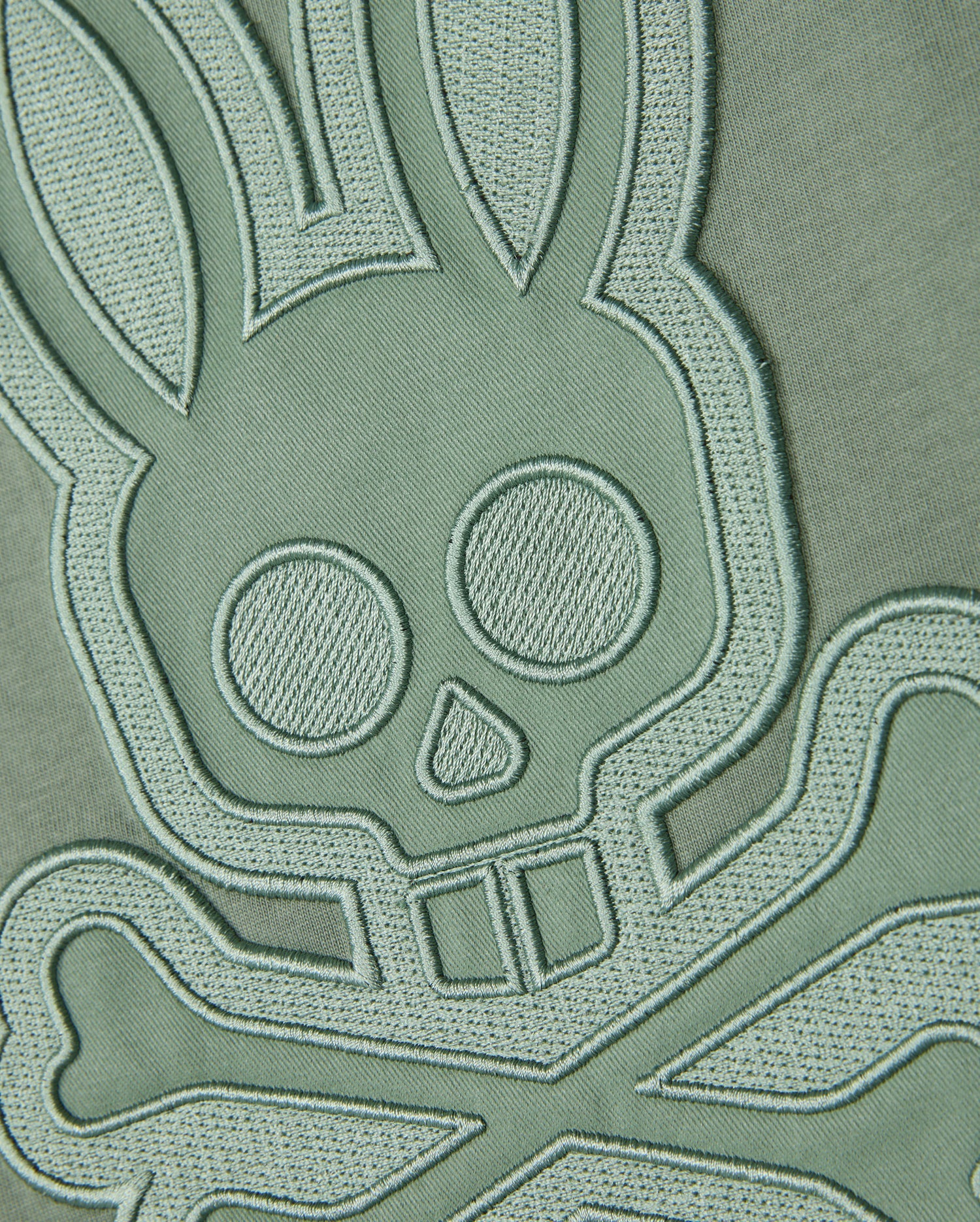A close-up of an embroidered patch on a Psycho Bunny MENS MALTON GRAPHIC TEE - B6U795D200 made from Pima cotton fabric. The design features a stylized skull with bunny ears and crossbones, detailed with stitching that raises it from the muted green background, giving it a three-dimensional look. Made in Peru, the unique appeal of this T-shirt is enhanced by its intricate craftsmanship.