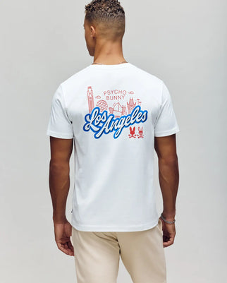 A person with short curly hair wears the MENS LOS ANGELES CITY TEE - B6U861A2PC by Psycho Bunny, featuring hand-drawn red graphics of city landmarks and "Psycho Bunny" in red with "Los Angeles" in blue script, paired with light-colored pants for a retro Dodgers vibe.