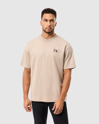 A man with short curly hair wears a Psycho Bunny MENS MONROE HEAVY WEIGHT RELAXED FIT TEE in beige and black pants, set against a plain gray background. The high-quality cotton tee features a small graphic on the left chest.