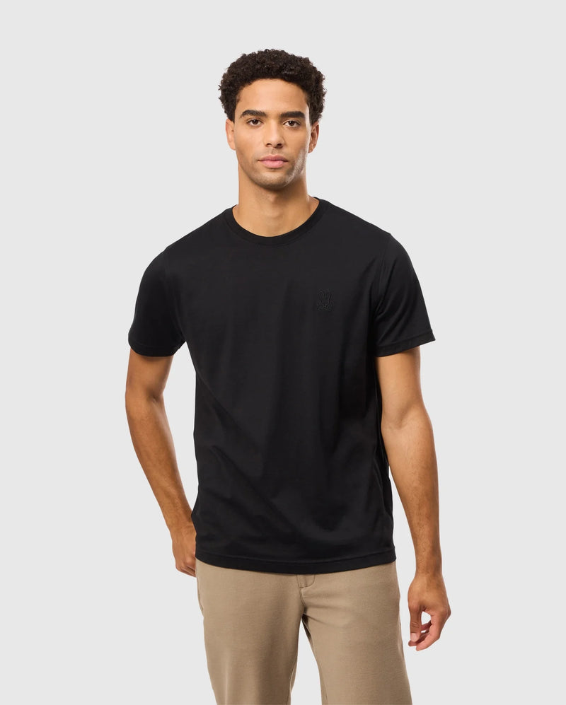 A man with short curly hair is standing against a plain light gray background, wearing the Psycho Bunny MENS JAMES PREMIUM CREW NECK TEE - B6U952D200, made of Pima cotton, and light brown pants. His left hand is in his pocket, and he has a neutral expression on his face.
