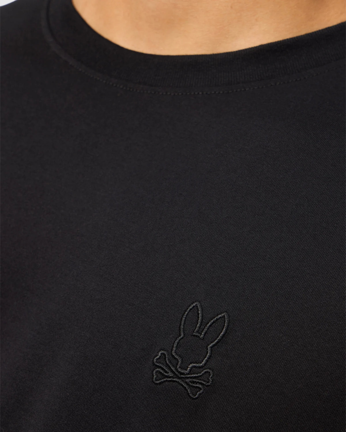 A close-up of a person showcasing the MENS JAMES PREMIUM CREW NECK TEE - B6U952D200 by Psycho Bunny. This black Pima cotton shirt features an embroidered black logo on the chest, depicting a bunny head with crossbones underneath. The person's neck and part of the shoulder are visible.