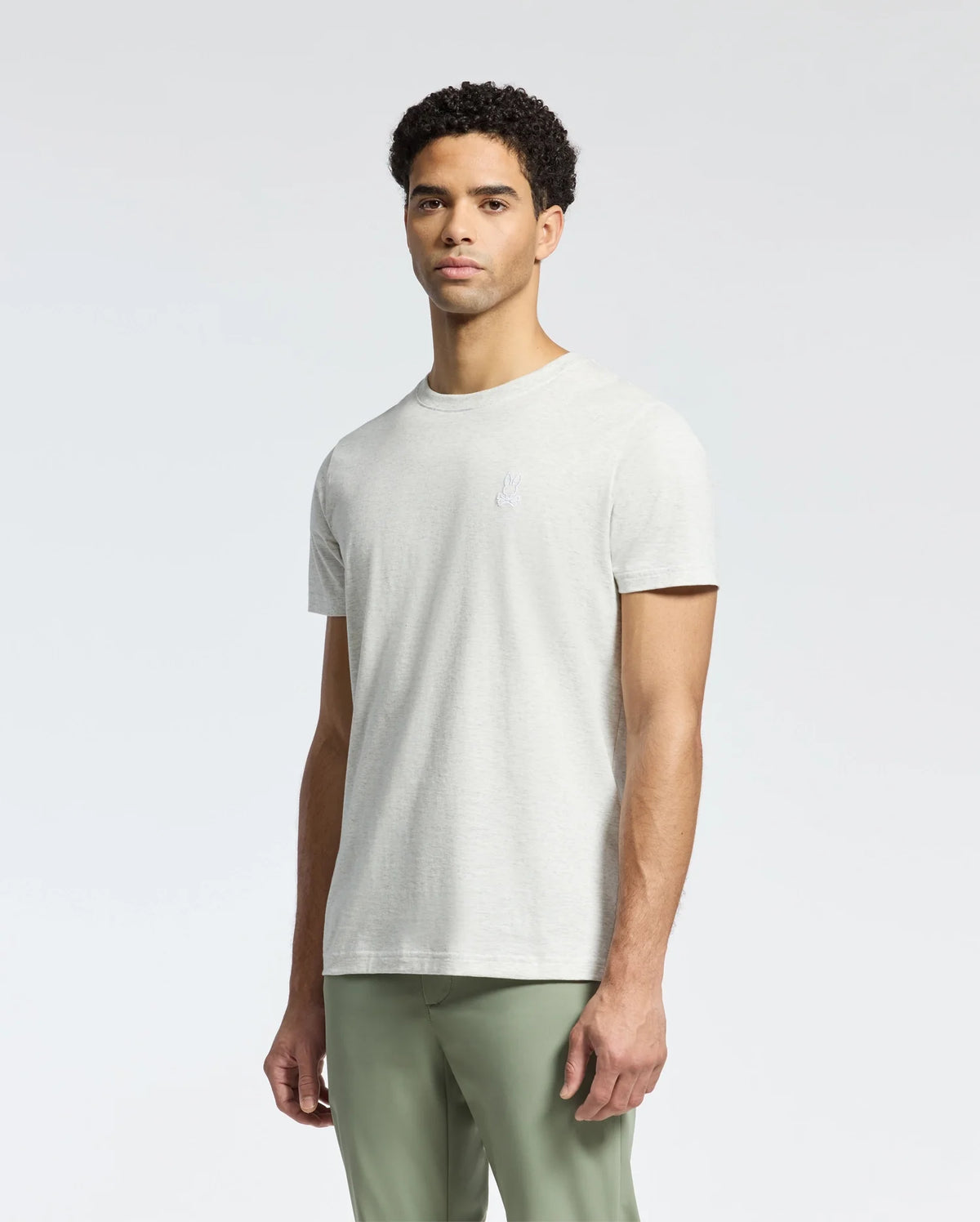 Wearing a Psycho Bunny MENS JAMES CREW NECK TEE in light gray, paired with light green pants, the person stands against a white background. With short, curly black hair and looking slightly off-camera, their attire highlights exquisite craftsmanship.