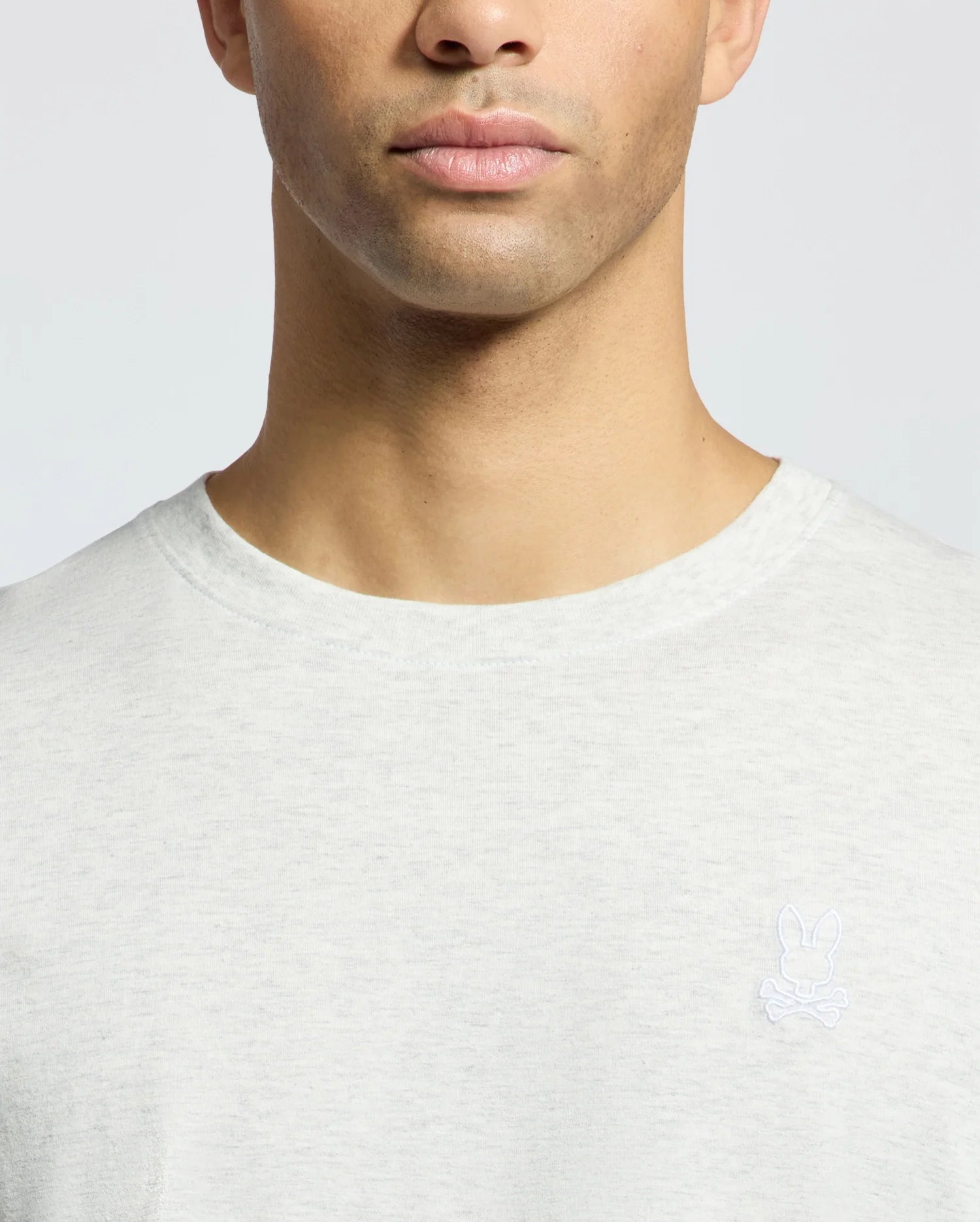 A person wearing the Psycho Bunny Men's James Crew Neck Tee in light gray, featuring a small white emblem of a rabbit above crossed bones on the left chest. The fine craftsmanship is shown, focused on the upper torso without fully revealing the face.