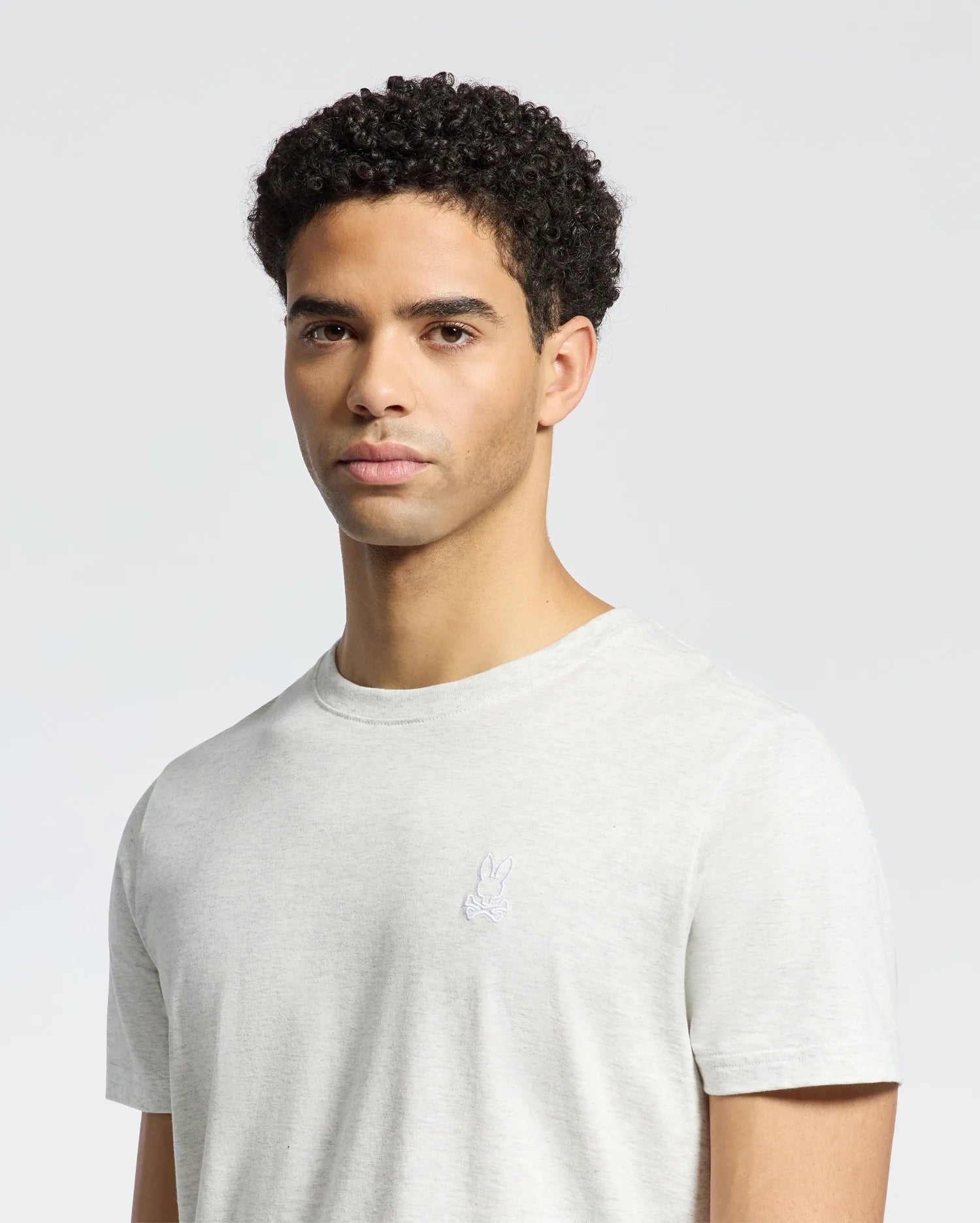 A person with short curly hair wears the Psycho Bunny Men's James Crew Neck Tee in light gray, crafted from luxurious Pima cotton with an embroidered small rabbit logo on the chest, against a plain white background.