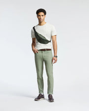 A man stands against a plain background wearing a white Psycho Bunny MENS JAMES CREW NECK TEE - B6U952D200, light green pants, and a black crossbody bag. He has short curly hair, finely crafted brown shoes, and is sporting a watch on his left wrist.