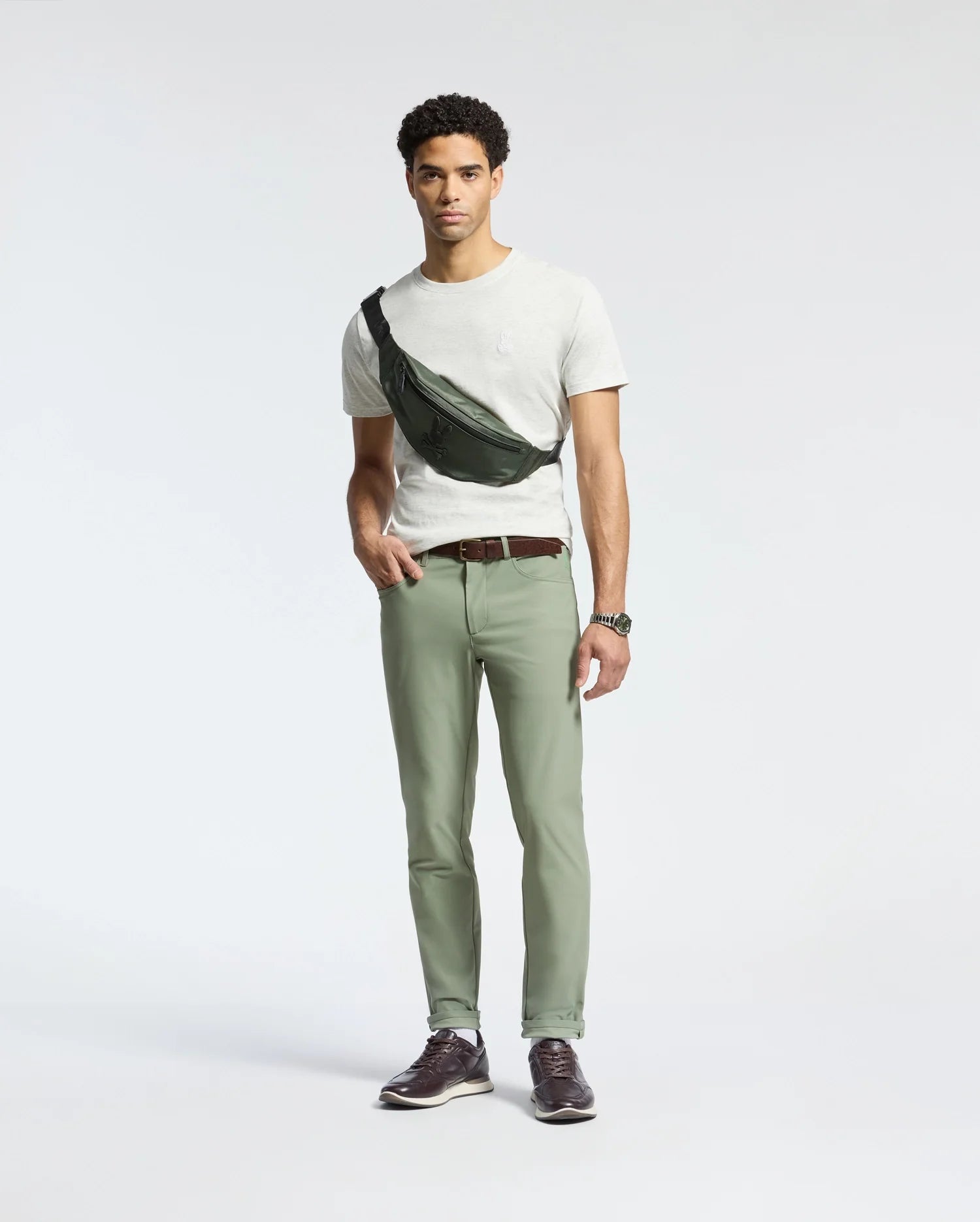 A man stands against a plain background wearing a white Psycho Bunny MENS JAMES CREW NECK TEE - B6U952D200, light green pants, and a black crossbody bag. He has short curly hair, finely crafted brown shoes, and is sporting a watch on his left wrist.