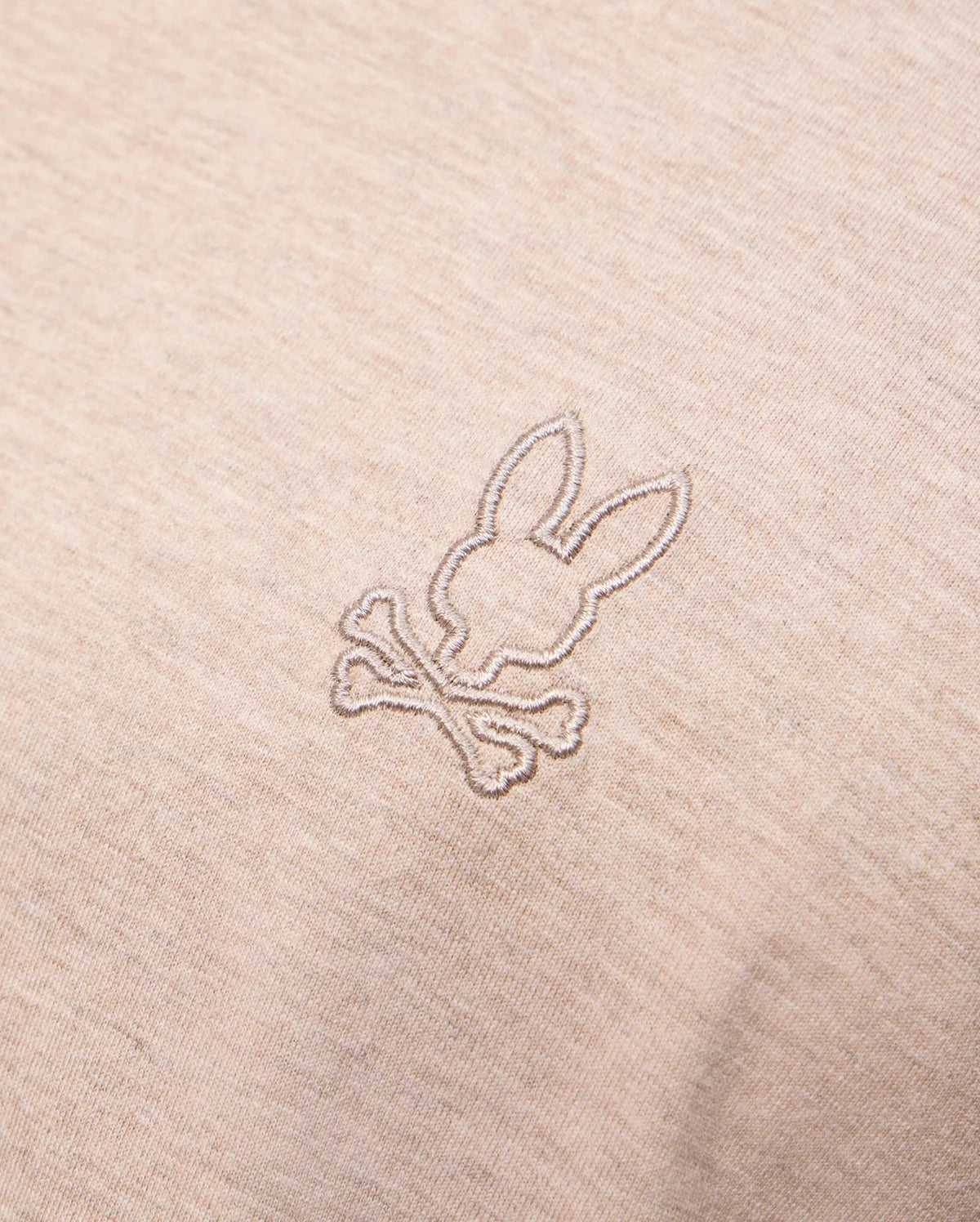 A close-up of the MENS JAMES PREMIUM CREW NECK TEE - B6U952D200 by Psycho Bunny in beige, crafted from premium Pima cotton, showcases a small, meticulously embroidered design featuring a stylized bunny head above two crossed bones, offering a playful twist on the classic skull and crossbones motif.