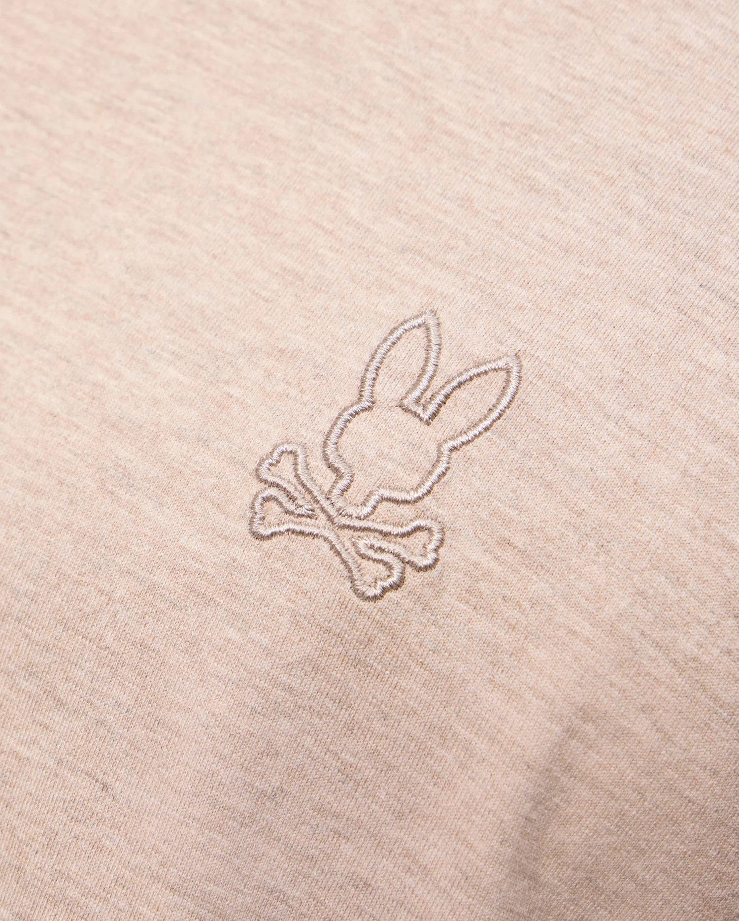 A close-up of the MENS JAMES PREMIUM CREW NECK TEE - B6U952D200 by Psycho Bunny in beige, crafted from premium Pima cotton, showcases a small, meticulously embroidered design featuring a stylized bunny head above two crossed bones, offering a playful twist on the classic skull and crossbones motif.