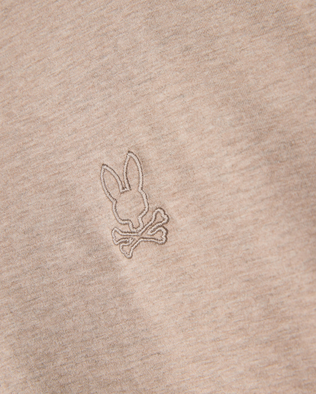 Close-up of the Psycho Bunny Men's James Premium Crew Neck Tee (B6U952D200) in premium Pima cotton, featuring an embroidered design of a bunny head on top of crossed bones, reminiscent of a skull and crossbones symbol. The meticulous embroidery is done in a slightly darker beige thread, offering a subtle contrast from the fabric.