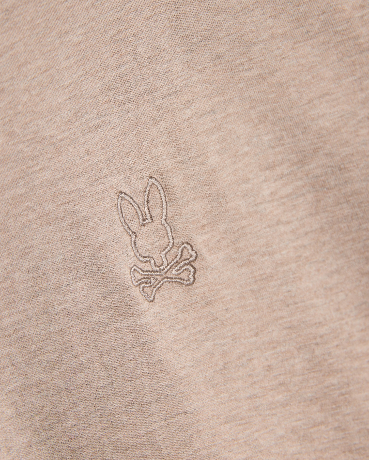 Close-up of the Psycho Bunny Men's James Premium Crew Neck Tee (B6U952D200) in premium Pima cotton, featuring an embroidered design of a bunny head on top of crossed bones, reminiscent of a skull and crossbones symbol. The meticulous embroidery is done in a slightly darker beige thread, offering a subtle contrast from the fabric.