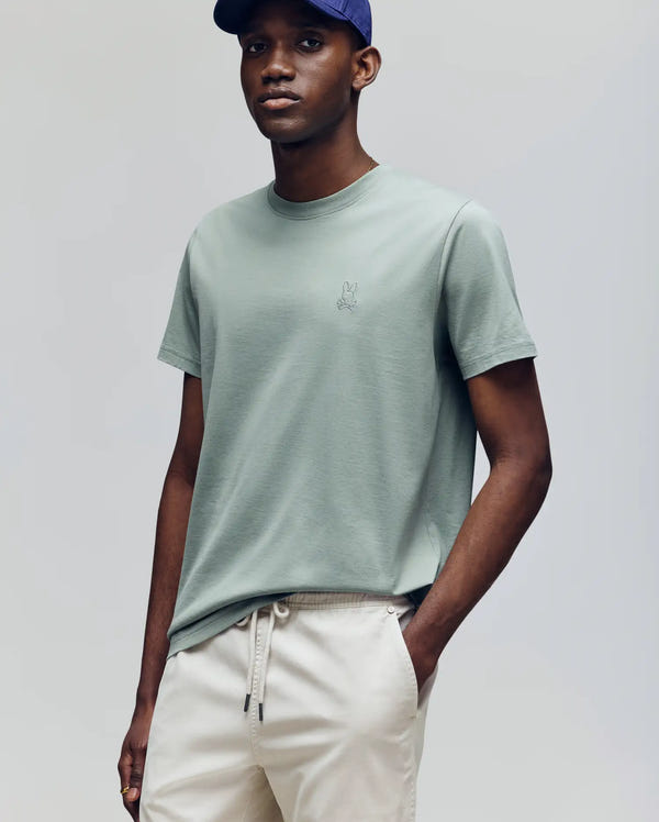 A man wearing the Psycho Bunny MENS JAMES PREMIUM CREW NECK TEE (B6U952D200) in light green with premium embroidery and beige drawstring pants stands with one hand in his pocket, also sporting a blue cap and looking to the side.